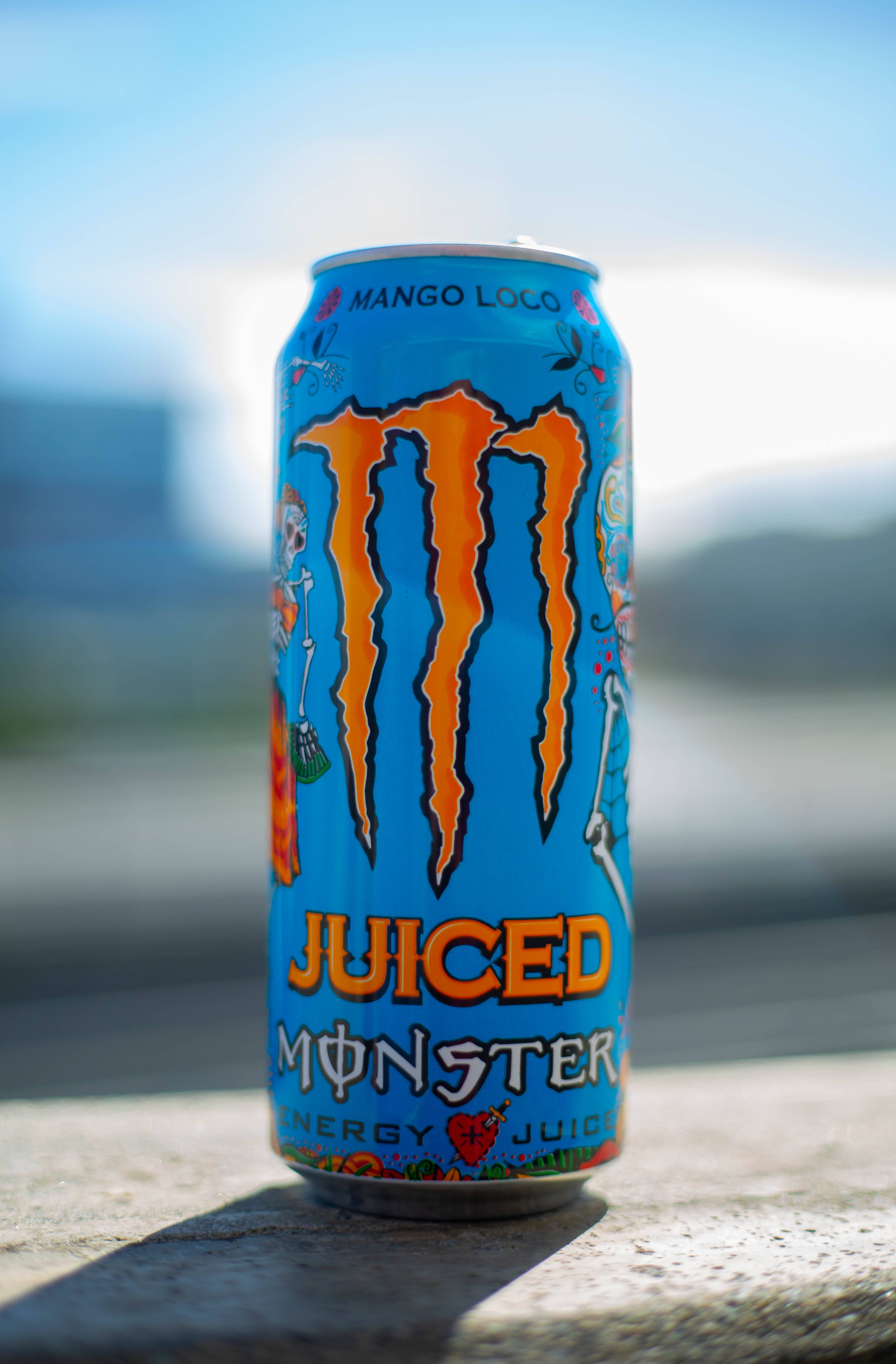 Juiced Monster Energy 電解質飲料罐 壁紙 — 一個充滿活力的藍色Monster Mango Loco能量飲料罐，帶有橙色爪標誌，放在陽光照射的混凝土表面上，背景是模糊的城市景觀。