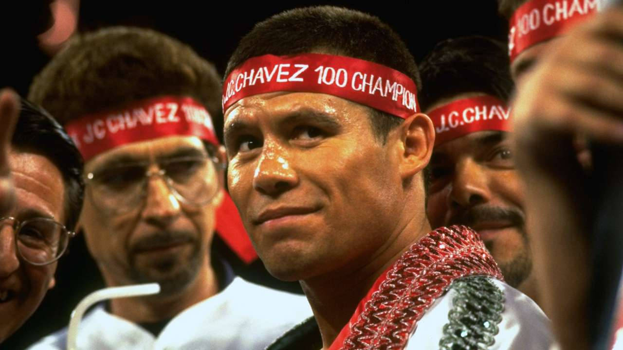 Julio César Chávez 獨特微笑 壁紙 — 一位戴著紅色 'J.C. CHAVEZ 100 CHAMPION' 頭帶的勝利拳擊手，在燈光昏暗的競技場中，於歡呼的粉絲間微笑。