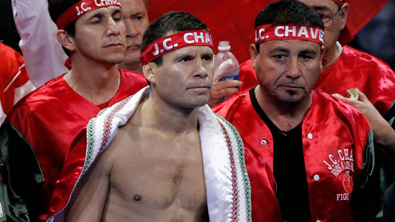 Julio César Chávez 穿袍子 壁紙 — 拳擊手J.C.查維斯Jr.赤裸上身，毛巾搭在肩上，被戴紅色頭帶和穿紅色外套的支持者包圍。