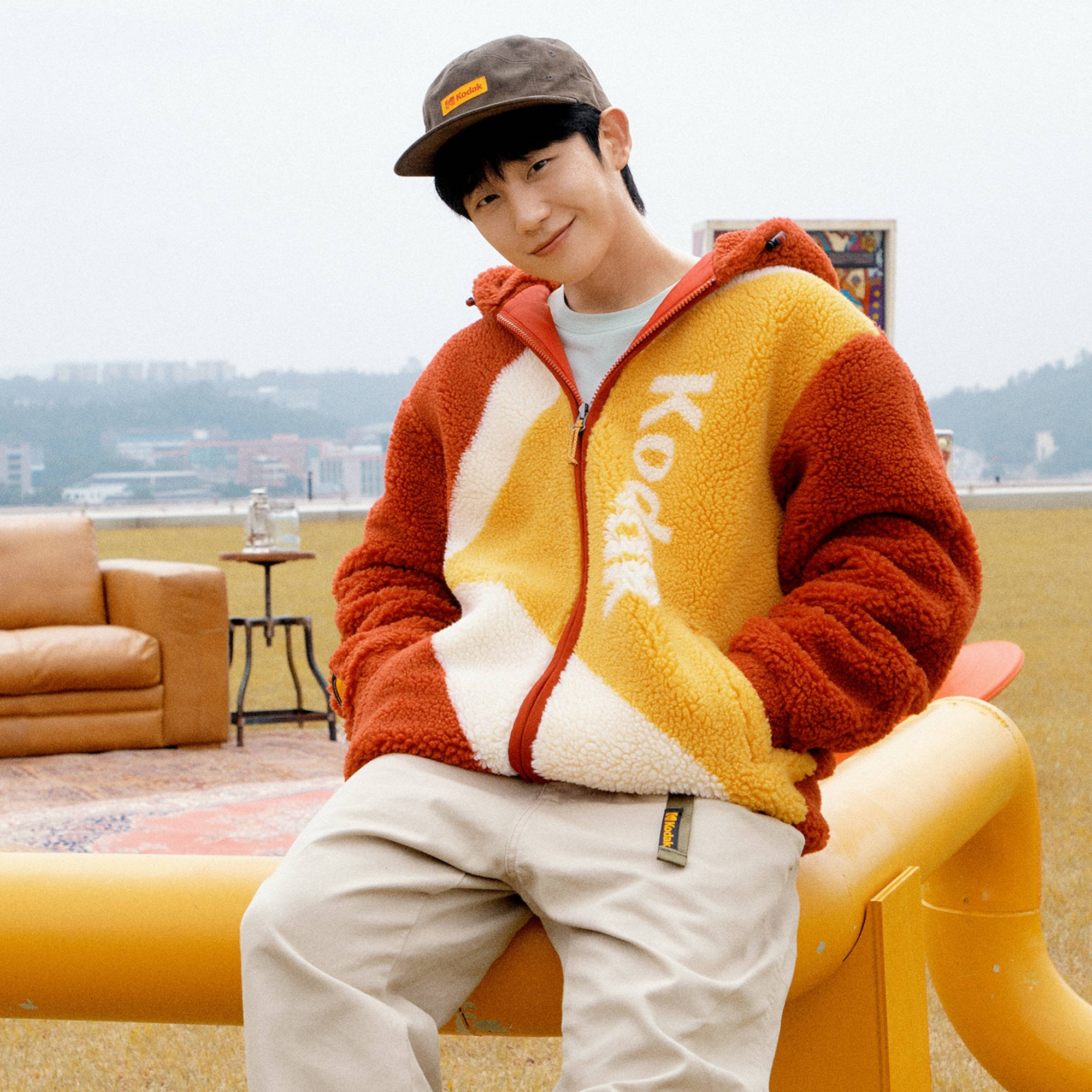 Jung Hae In Kodak微笑 壁紙 — 一個穿著彩色抓絨外套的年輕男子微笑著坐在一張明亮的黃色長椅上，俯瞰城市景觀。
