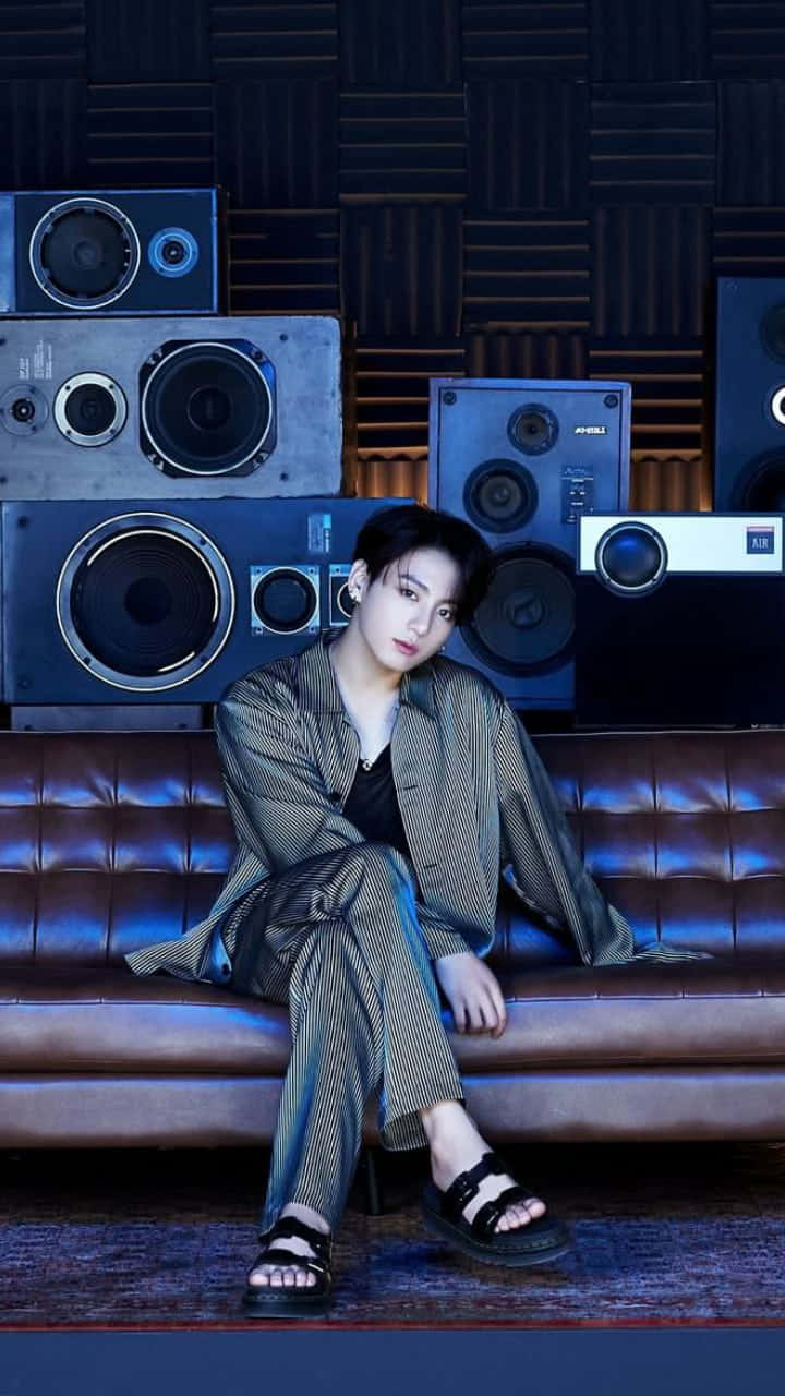 Jungkook 展現時尚造型 壁紙 — 一位穿著條紋睡衣的年輕男子，在燈光昏暗的工作室裡，放鬆地坐在一張簇絨皮革沙發上，周圍環繞著復古喇叭。