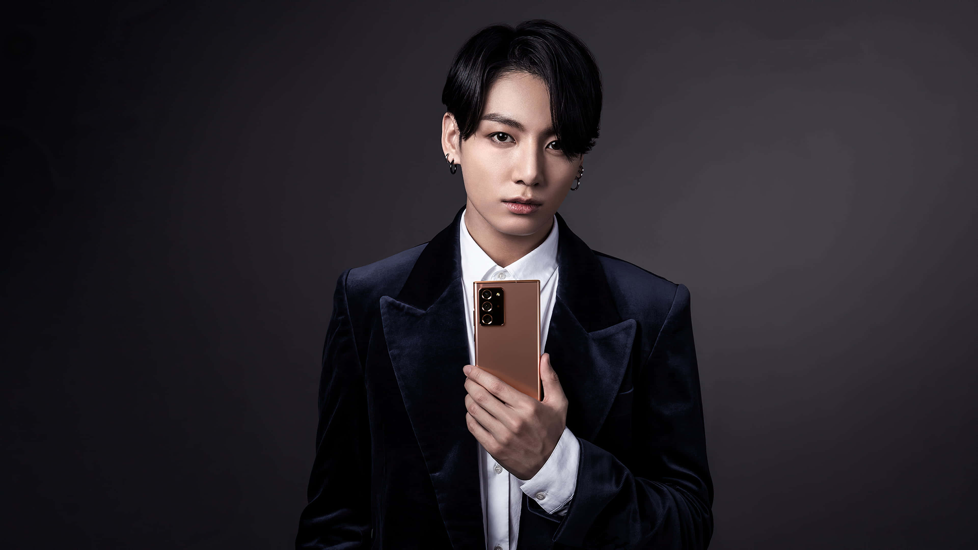 Jungkook 穿白色毛衣微笑 壁紙 — 一名身穿深色天鵝絨西裝的男子，在陰鬱的灰色背景下手持玫瑰金智能手機，目光直接而強烈。