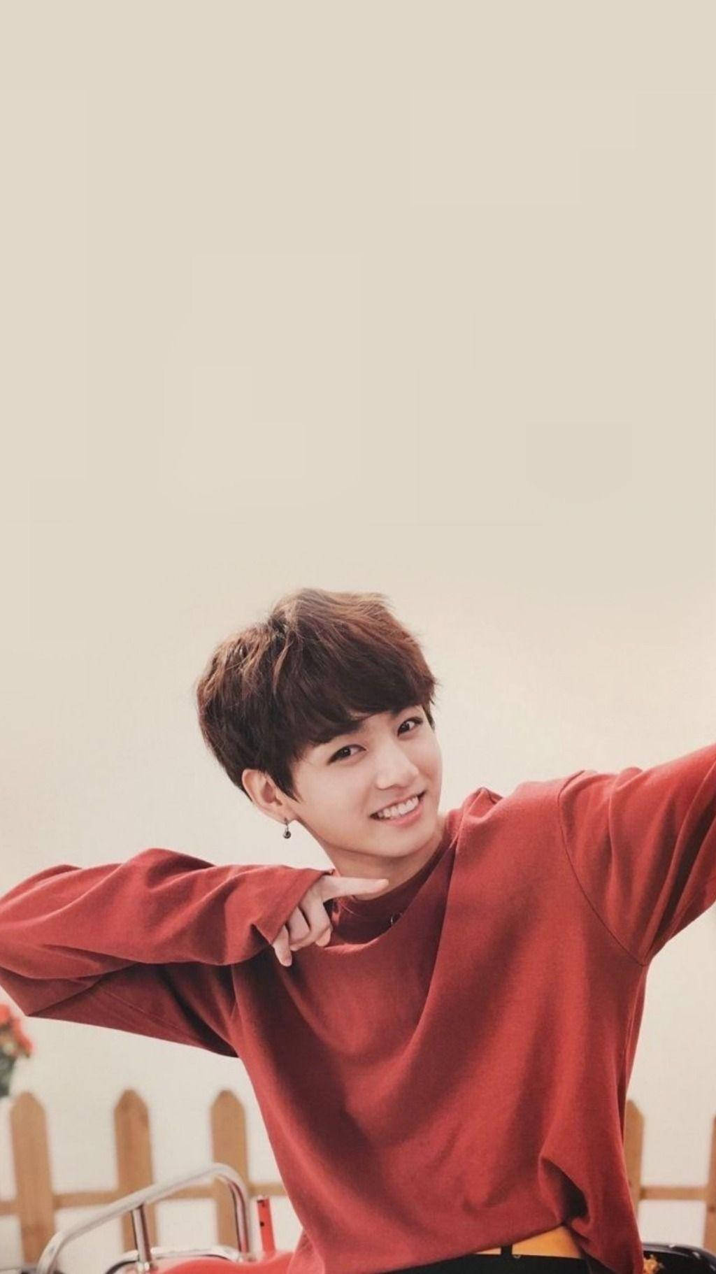Jungkook 美學熱潮姿勢 壁紙 — 一位身穿紅色毛衣的年輕男子，在柔和的米色背景前張開雙臂，露出燦爛的笑容。