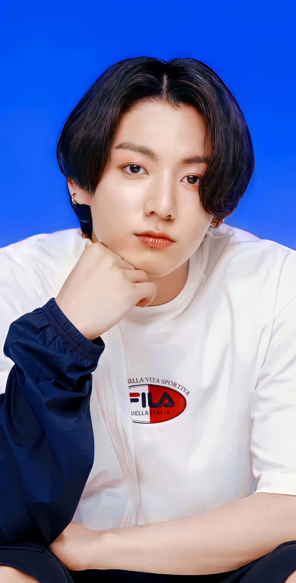 Jungkook以迷人眼神征服觀眾 壁紙 — 一位深色頭髮的年輕男子，在純藍色背景前，穿著白色FILA襯衫，將下巴擱在手上。