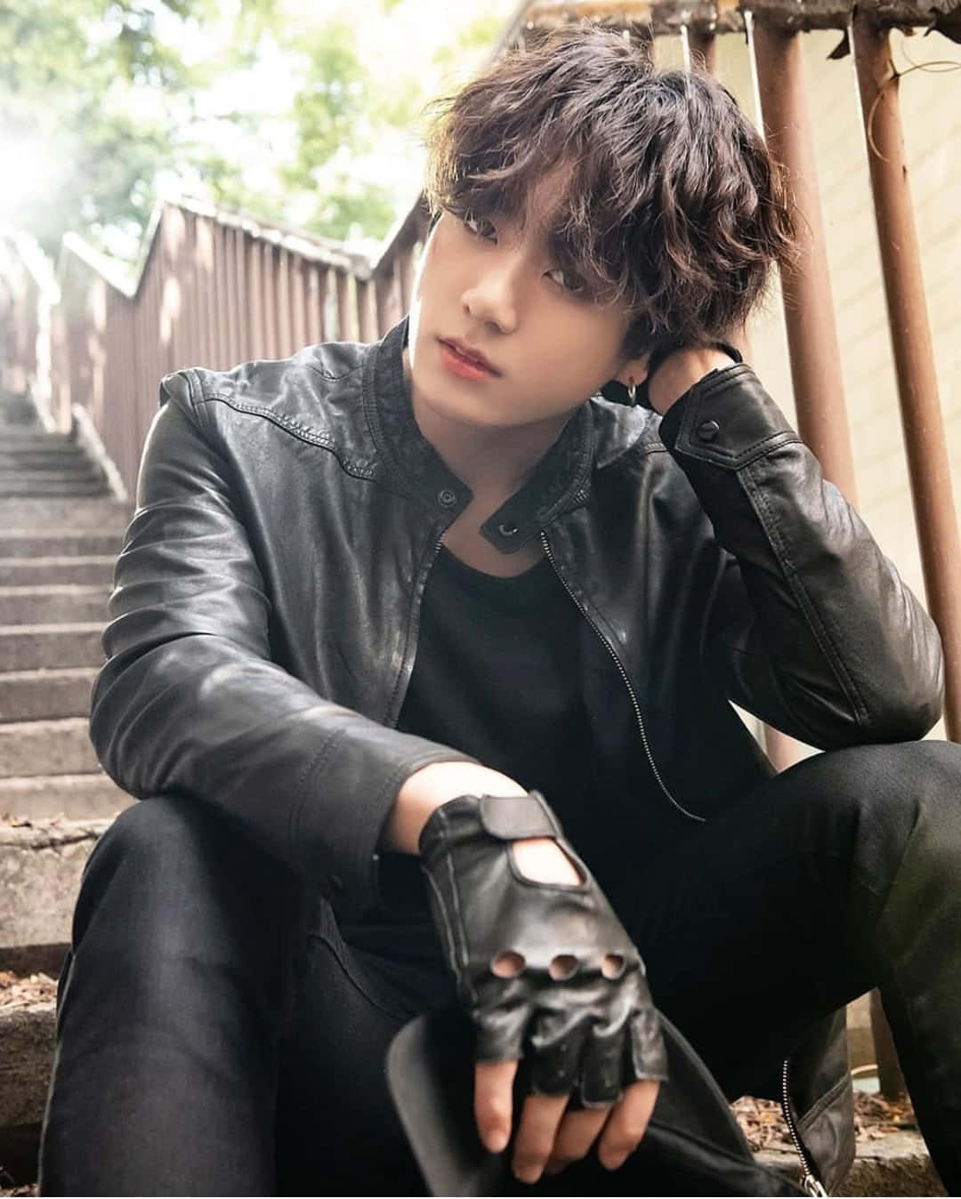 Jungkook在近照中溫暖微笑 壁紙 — 一個頭髮凌亂的年輕男子穿著黑色皮褸和露指手套坐在石階上，若有所思地向上凝視。