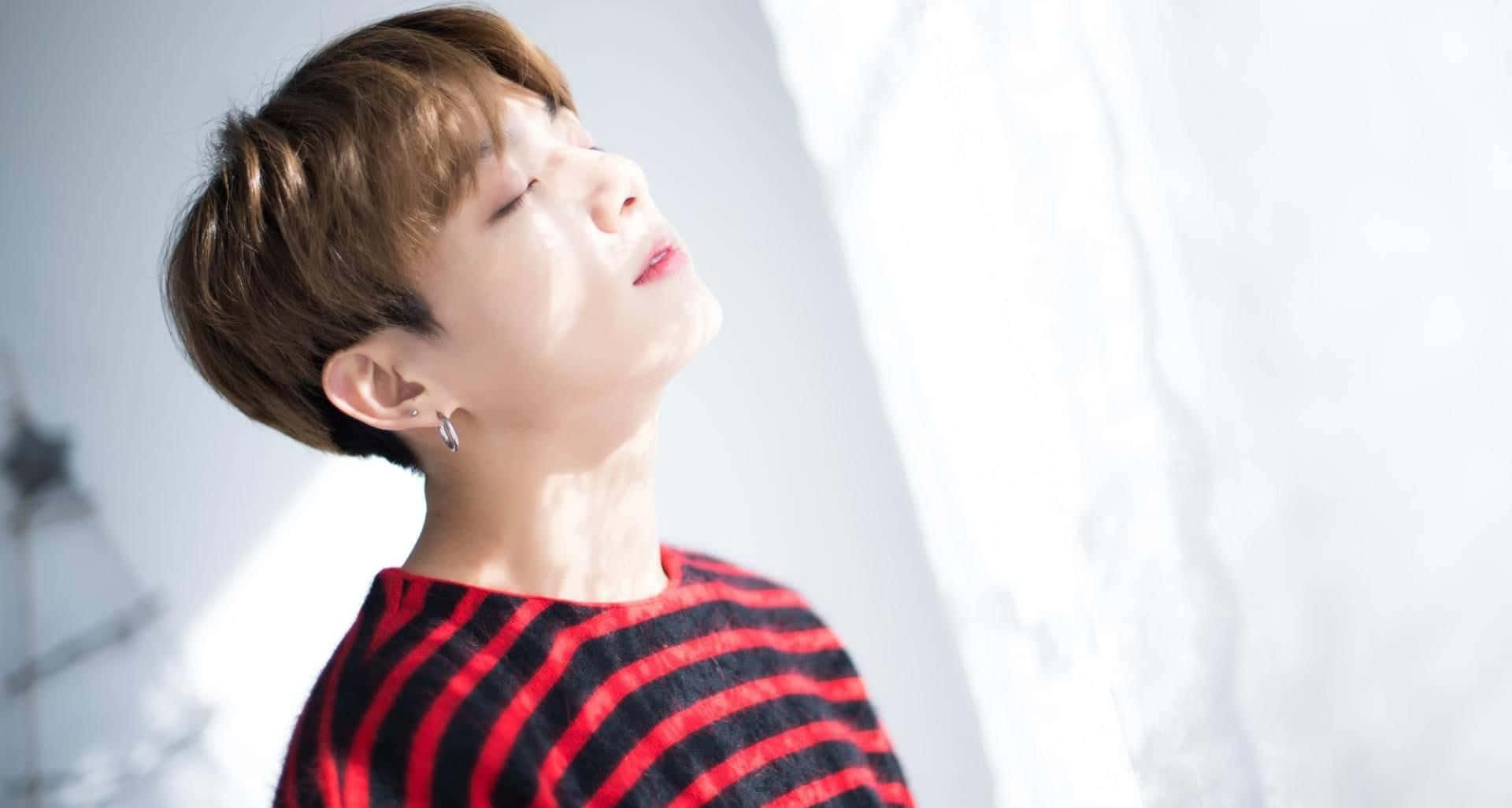 Jungkook擁抱熱情粉絲 壁紙 — 一位短棕髮、身穿紅條紋毛衣的年輕人在窗邊柔和的陽光下向後仰頭。