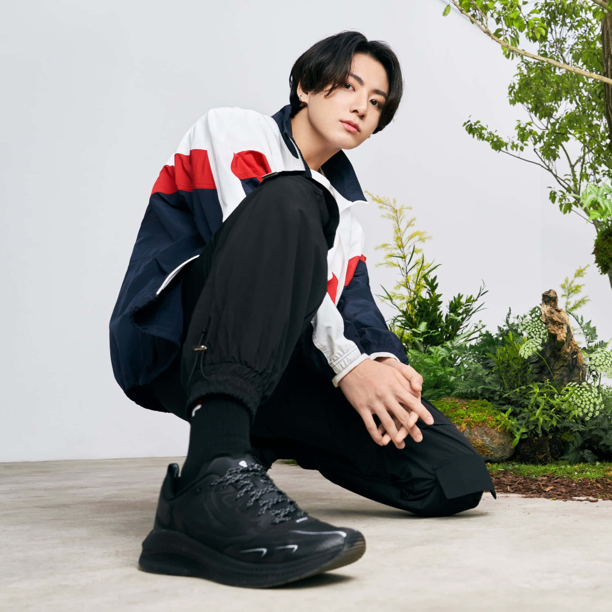 Jungkook於休閒姿態中展現燦爛笑容 壁紙 — 一名身穿彩色運動外套的年輕男子蹲在茂密綠植旁，背景是純白牆壁。