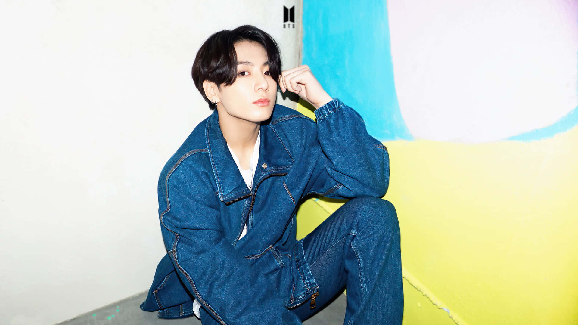 Jungkook於煙霧背景中展現時尚姿態 壁紙 — 一位身穿牛仔裝的年輕男子隨意地坐在色彩繽紛的牆邊，牆上有大膽的黃色和藍色區域。