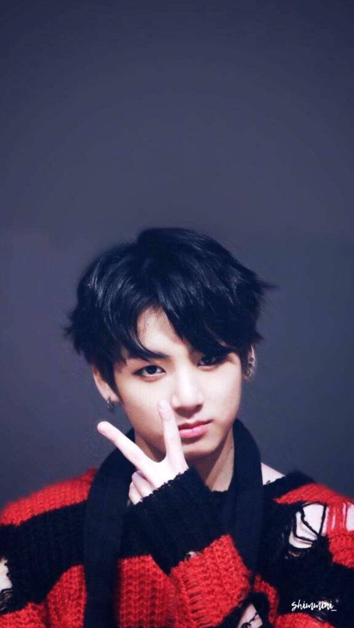 Jungkook時尚V字造型 壁紙 — 一位黑髮、眼神銳利的年輕男子穿著紅黑針織毛衣，在柔和的灰色背景前做出和平手勢。