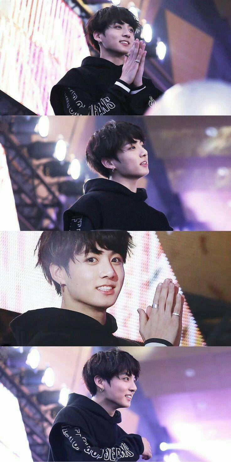 Jungkook時尚黑色連帽衫 壁紙 — 一名身穿黑色連帽衫的年輕男子在燈光璀璨的舞台上微笑並鼓掌，身後是五彩繽紛的燈光和設備。