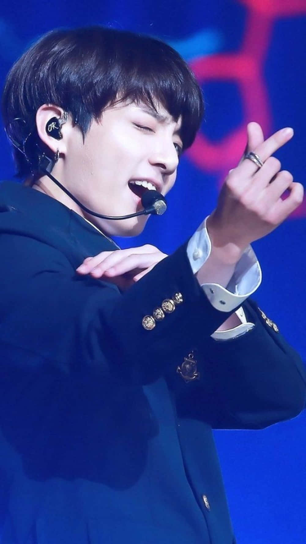 Jungkook眨眼BTS手指心形 壁紙 — 一位男性表演者在藍色舞台燈光下穿著深色西裝外套熱情地唱歌。