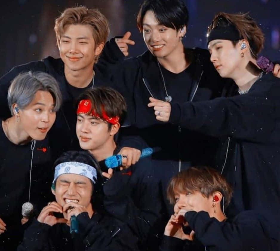 Jungkook神情深思，卻依然自信滿滿 壁紙 — 七位男K-pop偶像身穿黑色服裝，在明亮的燈光下，手持麥克風和彩色頭帶，在舞台上俏皮地擺姿勢。