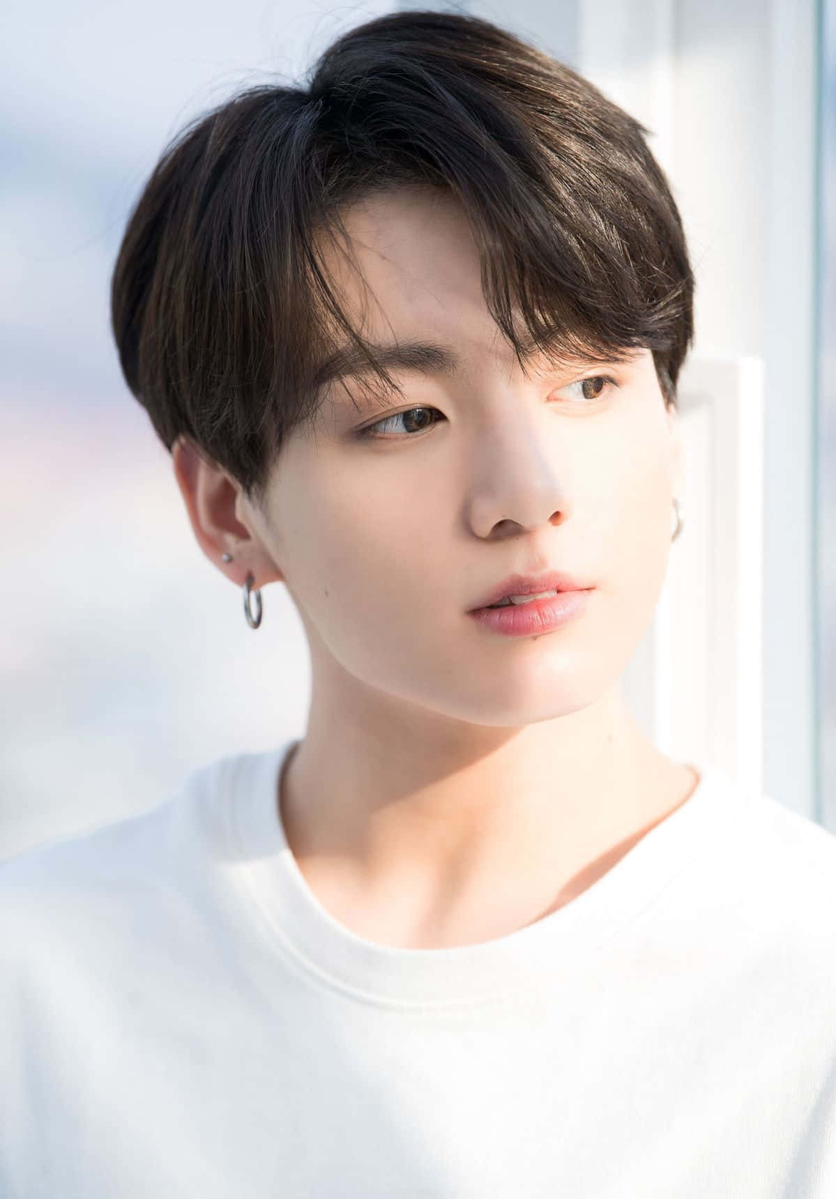 Jungkook穿休閒服裝微笑 壁紙 — 一位黑髮戴著圈狀耳環的年輕男子，穿著白色T恤，在明亮的窗戶背景下若有所思地凝視著。