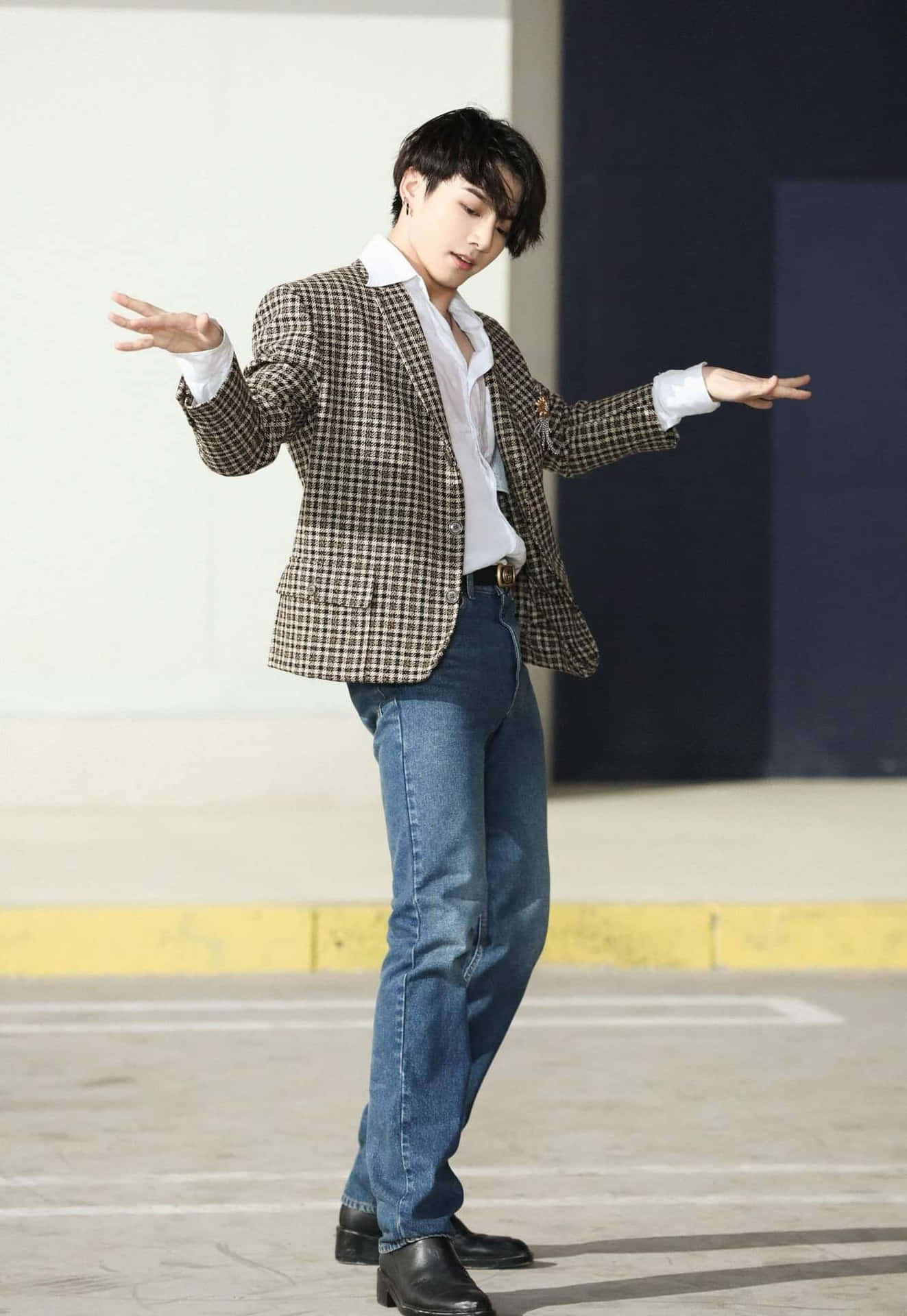 Jungkook舞台激情演出 壁紙 — 一位身穿格仔西裝外套和牛仔褲的年輕男子，在極簡城市背景下伸展雙臂，擺出充滿動感的姿勢。