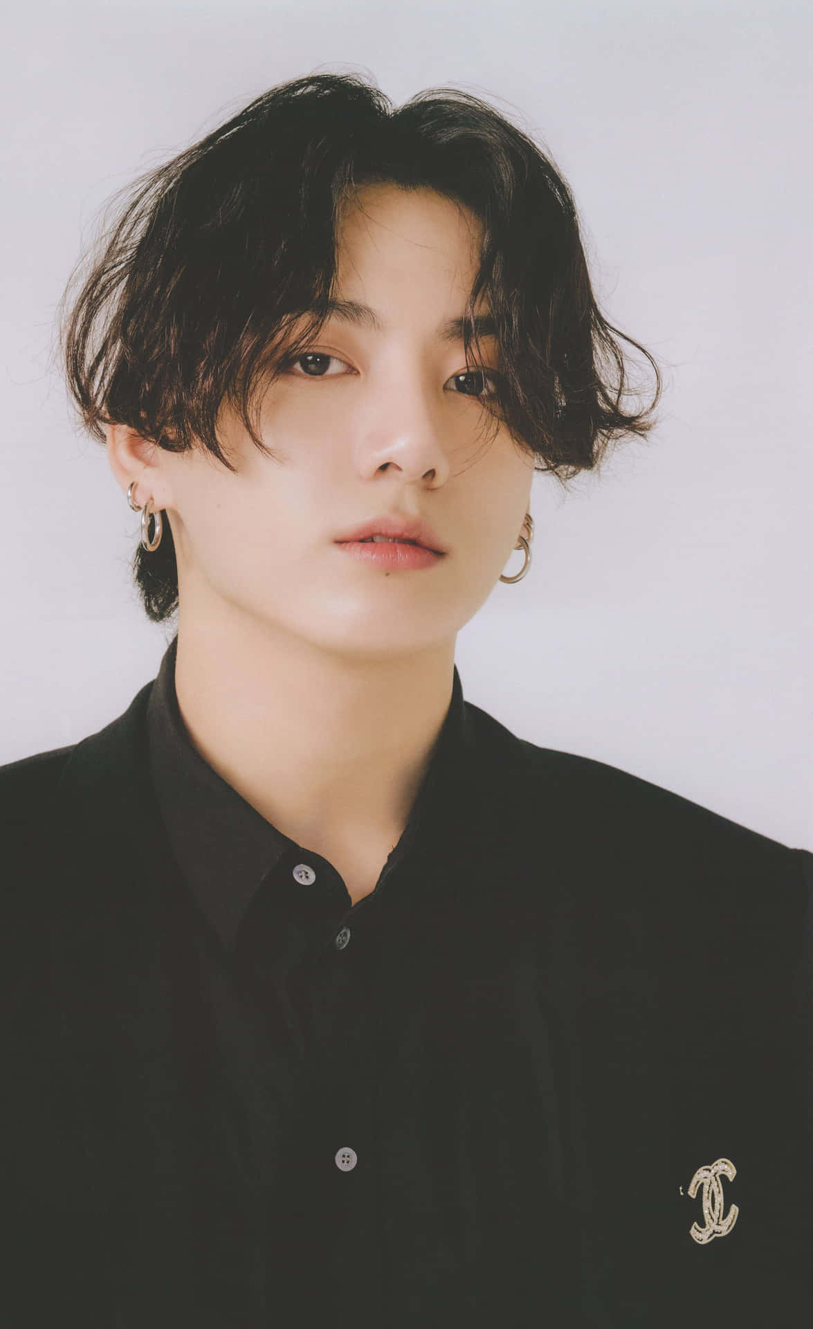 Jungkook舞台精彩瞬間 壁紙 — 一位深色波浪髮、戴金色圓環耳環的年輕男子，身穿黑色鈕扣襯衫，別著Chanel標誌胸針，背景為純白色。