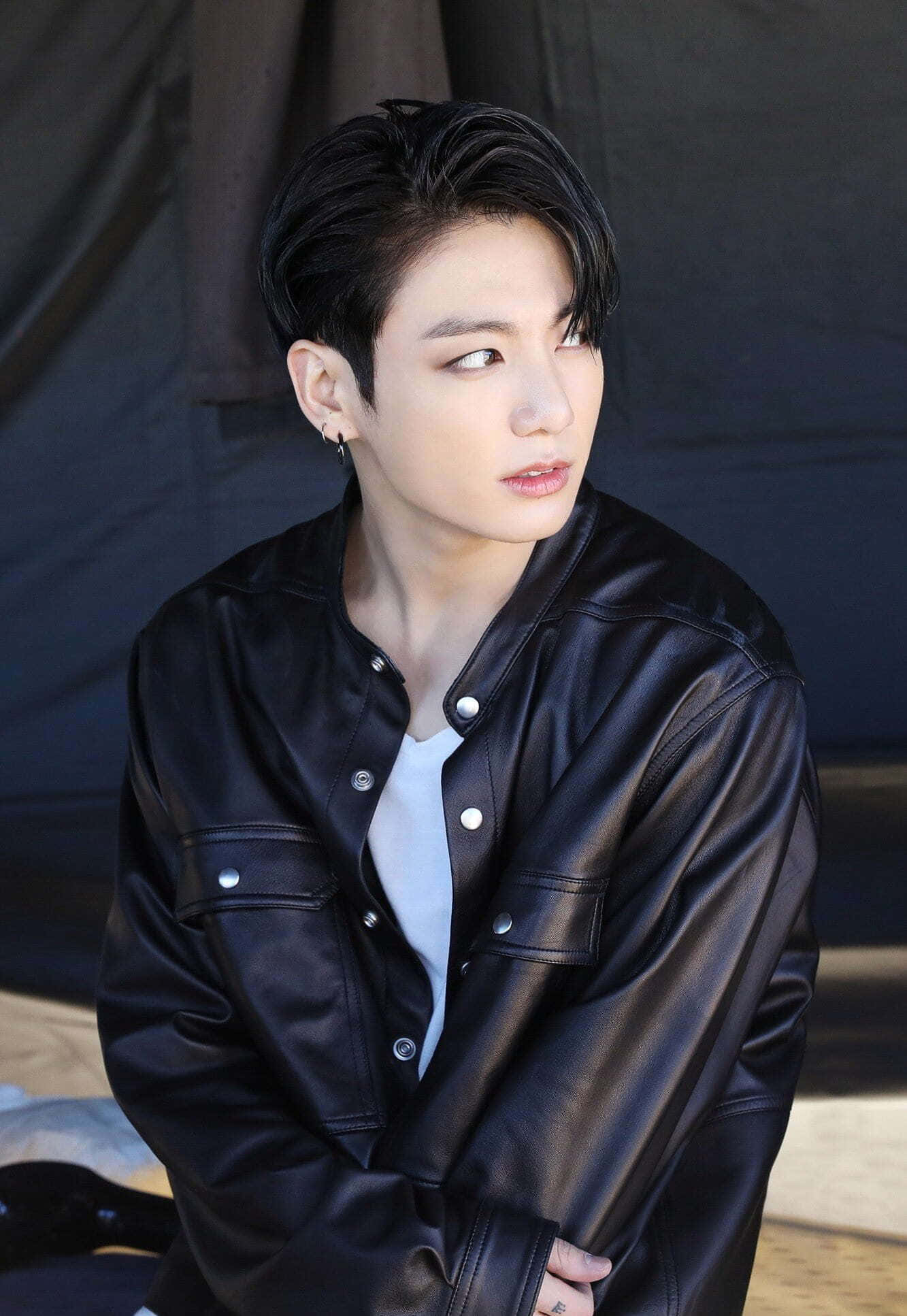 Jungkook藍色服裝微笑時刻 壁紙 — 一位深色頭髮的年輕男子穿著黑色皮褸內搭白色T恤，在深色背景下側目而視。