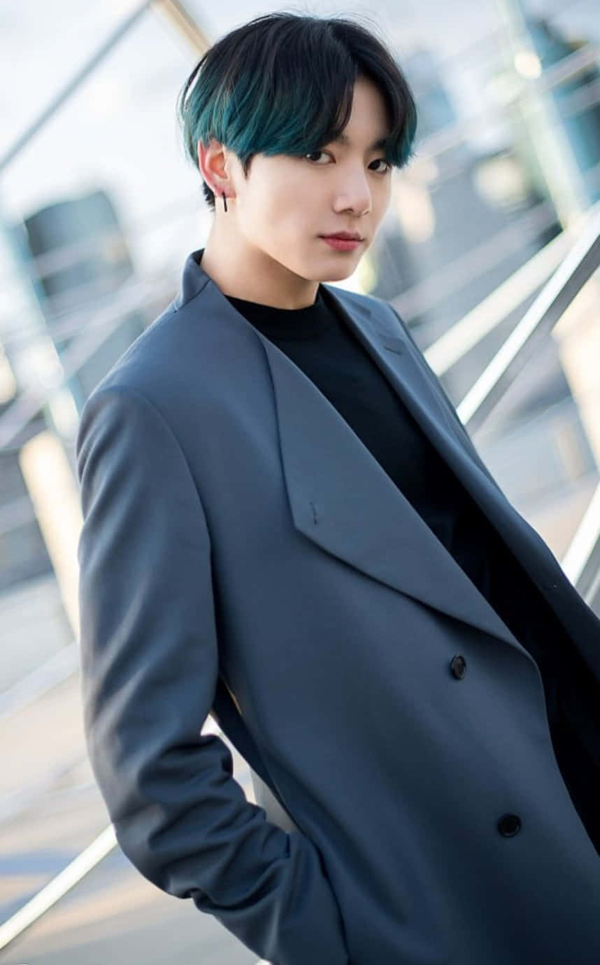 Jungkook迷人近距離肖像 壁紙 — 一位年輕男子，帶有藍綠色髮尾和灰色外套，自信地站在模糊的現代玻璃建築背景前。