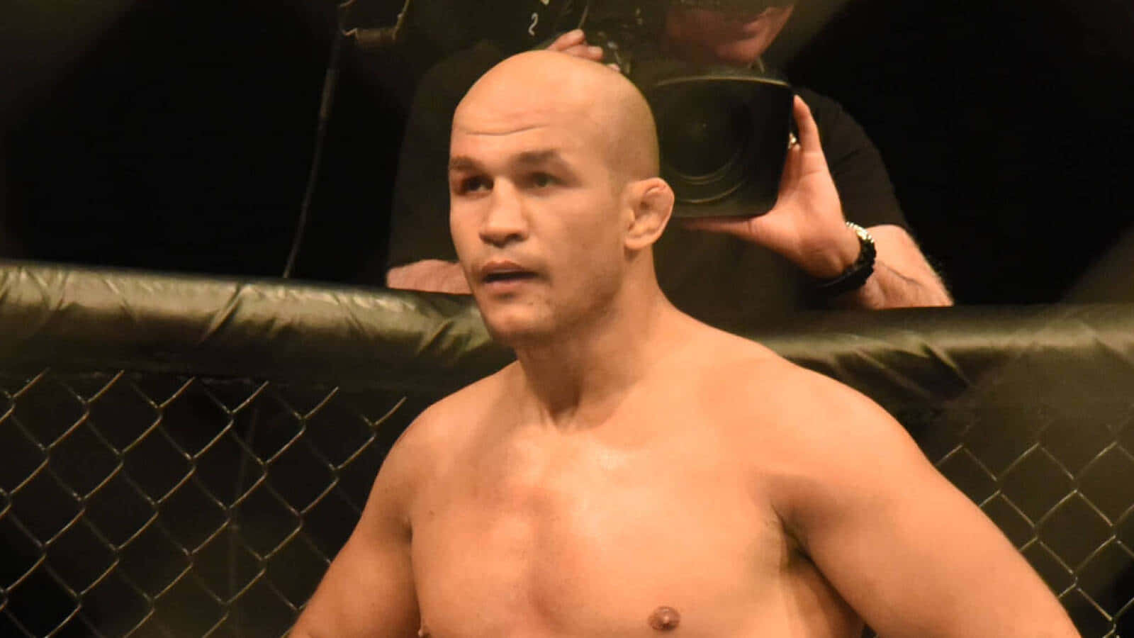 一位光頭MMA戰士在黑色八角籠內，身後有一名攝影師。