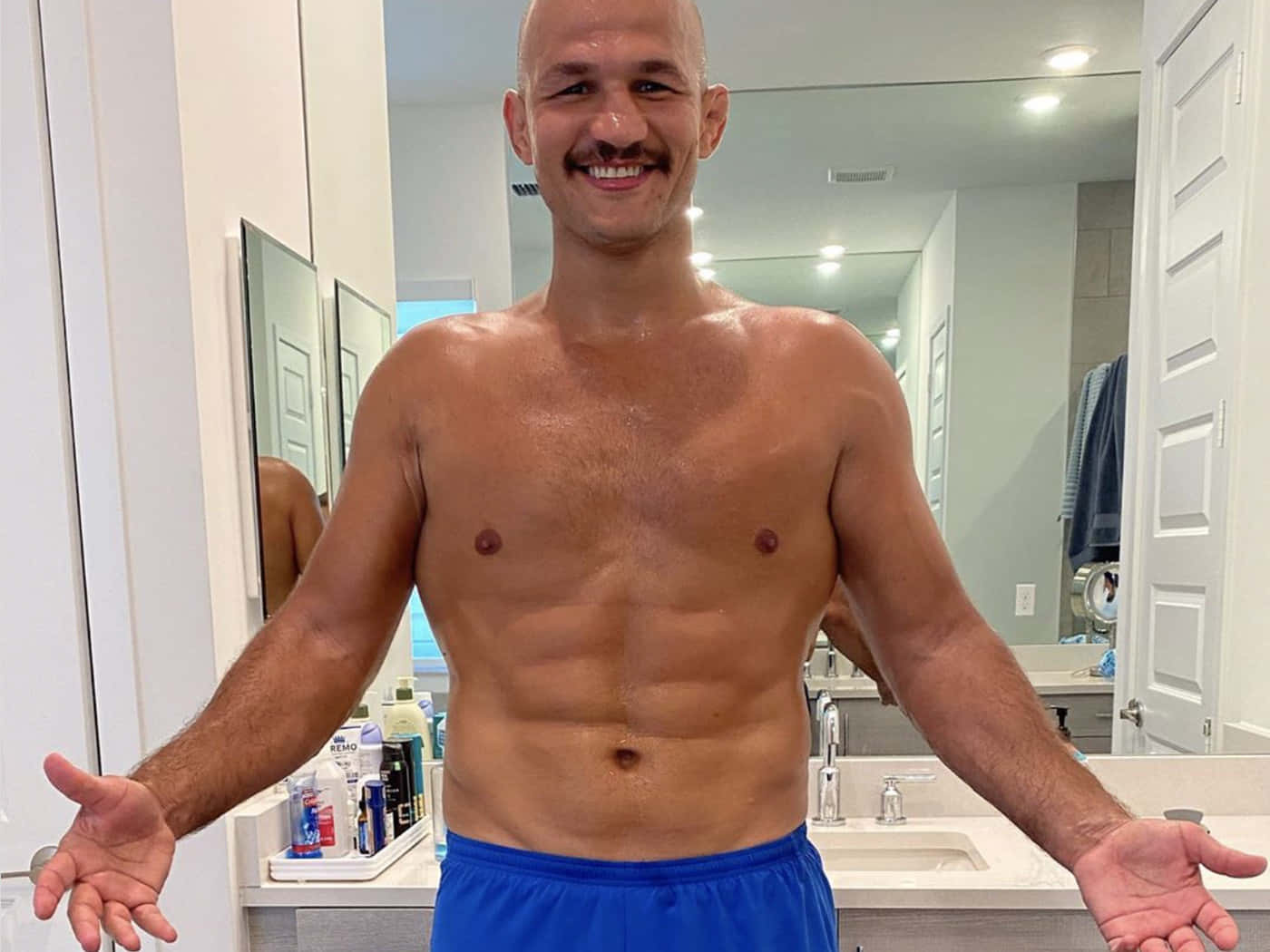 Junior Dos Santos 展現減重成果 壁紙 — 一位留鬍鬚、微笑的肌肉男赤裸上身站在明亮的浴室，雙臂張開，展示他結實的身材。