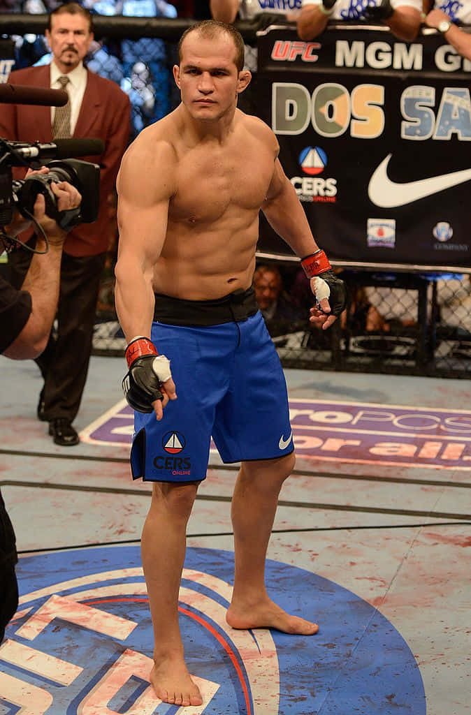 Junior Dos Santos 在 MGM Grand Garden Arena 對戰 Cain Velasquez 壁紙 — 一位肌肉發達的MMA拳手穿著藍色短褲，在UFC賽事中赤腳站在沾滿血跡的八角籠中央。