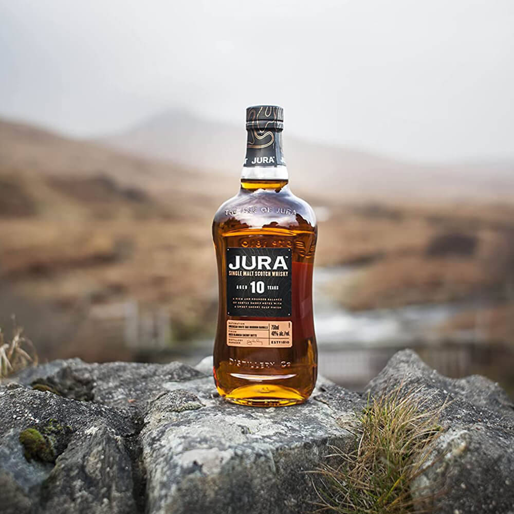 Jura 18年陳釀麥芽威士忌 壁紙 — 一瓶Jura 10年單一麥芽威士忌放在粗糙的灰色岩石上，背景是霧氣瀰漫的山丘。