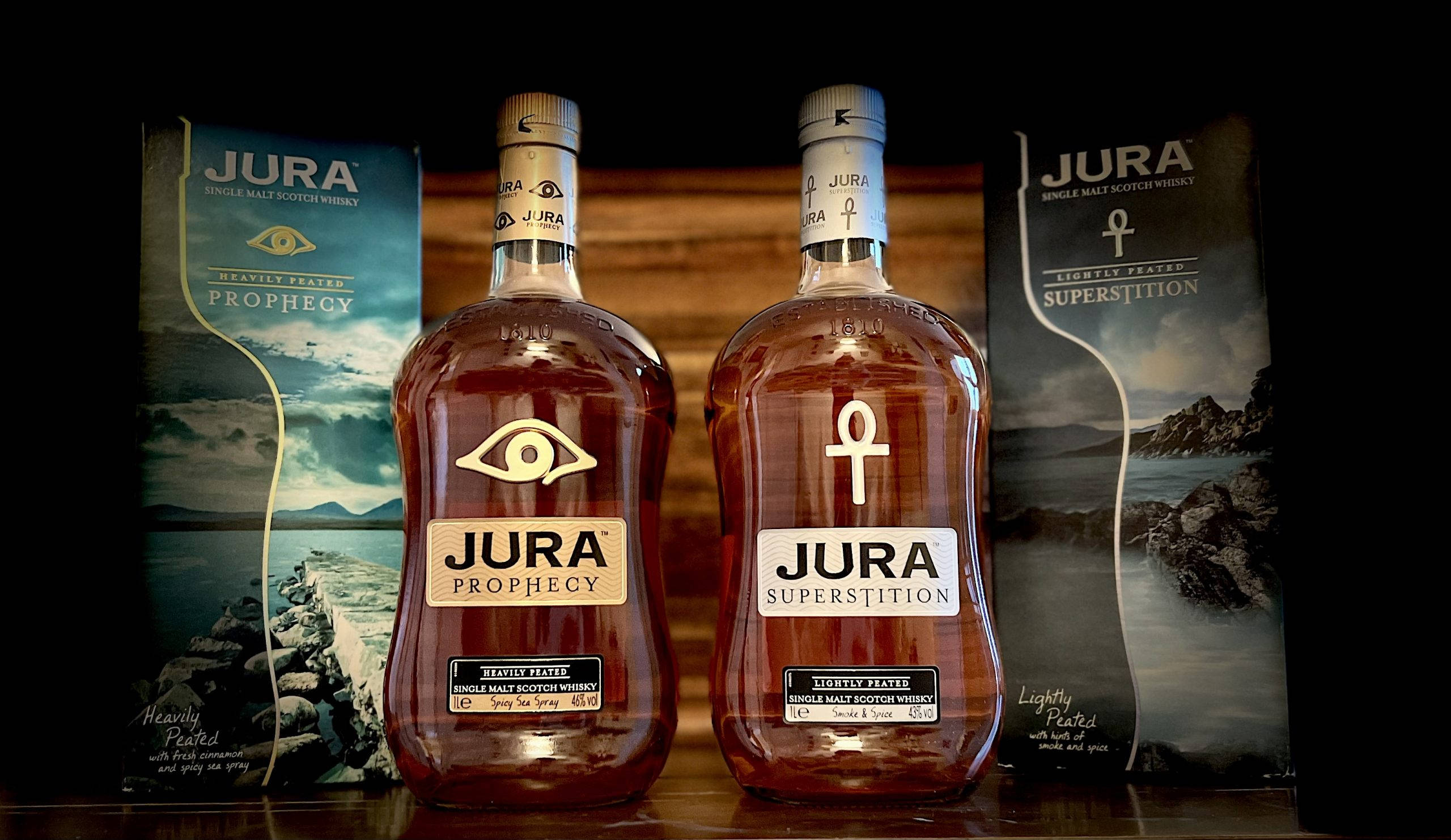 Jura 島 Superstition 威士忌 壁紙 — 兩瓶Jura威士忌，標籤為Prophecy和Superstition，在深色木質背景前被照亮，後面有品牌盒子。