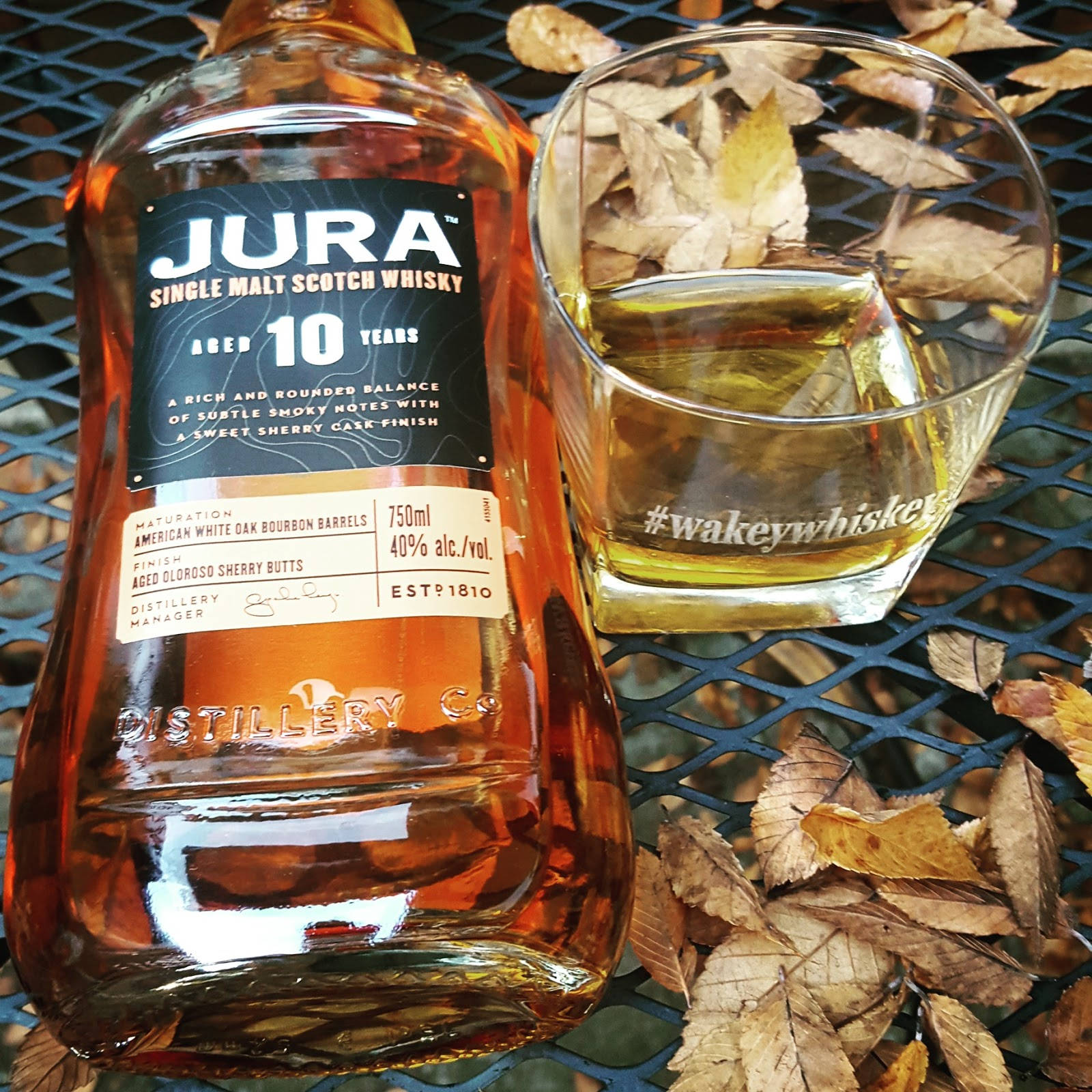 Jura最上乘威士忌烈酒 壁紙 — 一瓶Jura 10年單一麥芽蘇格蘭威士忌放在一個金屬網桌上，旁邊有一個裝滿琥珀色液體的玻璃杯，周圍環繞著乾燥的秋葉。