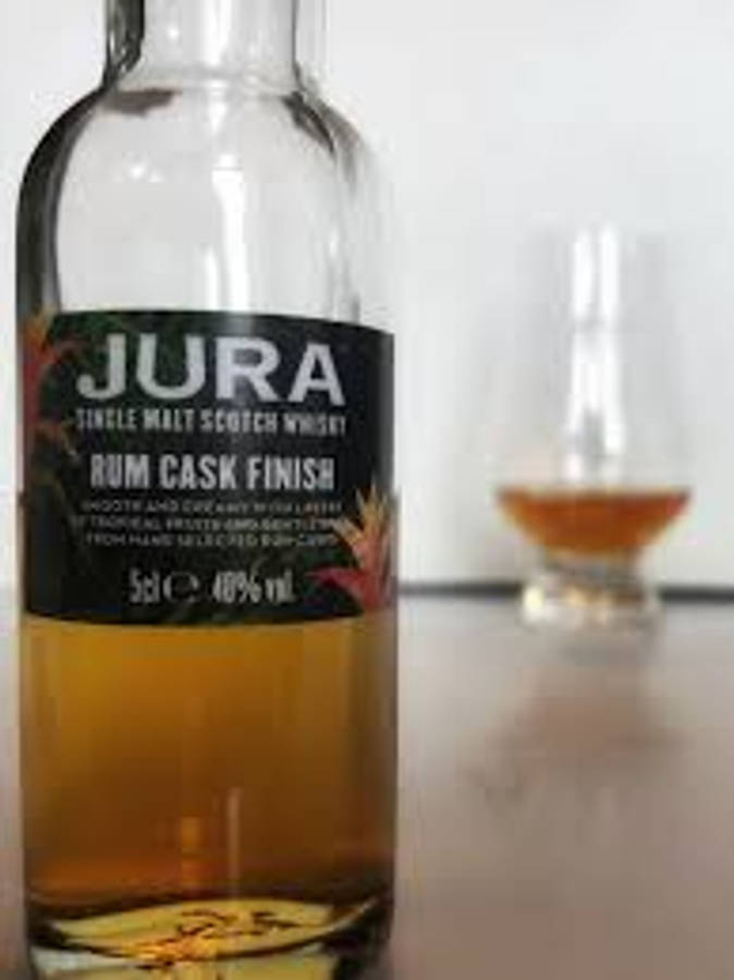 Jura朗姆桶陳威士忌 壁紙 — 一瓶清晰的Jura Rum Cask Finish威士忌在焦點中，背景中有一杯琥珀色液體模糊不清。