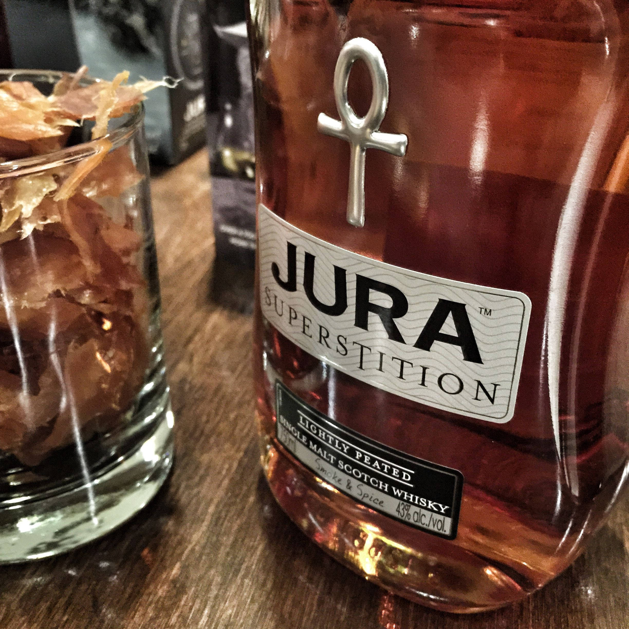 Jura超級迷信威士忌 壁紙 — Jura Superstition 威士忌酒瓶的特寫，帶有安卡標誌，旁邊係一個裝有乾木屑嘅玻璃杯，放喺木枱上。