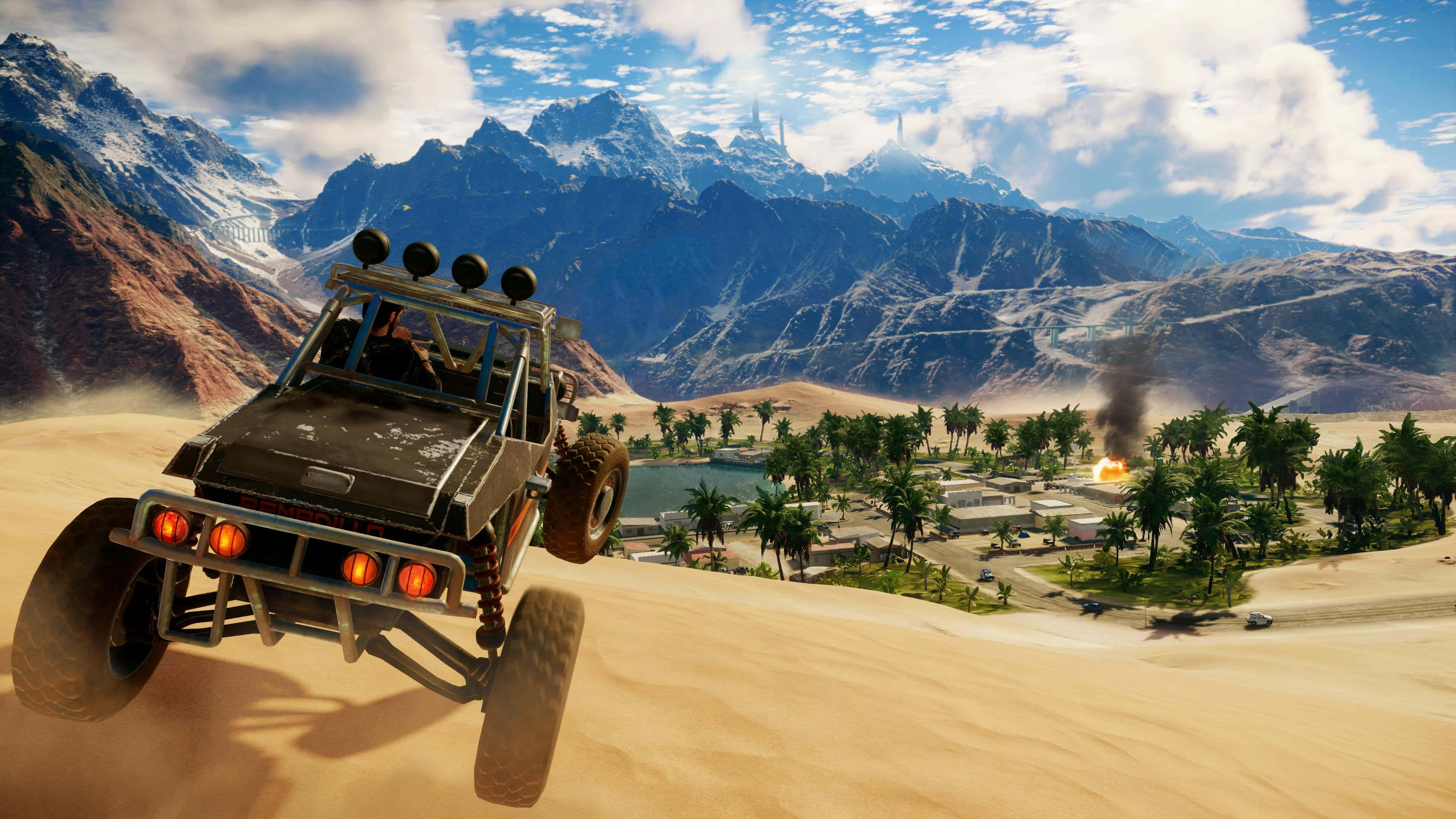 《Just Cause 4》：行動即將到來 壁紙 — 一輛堅固的越野車飛越沙丘，朝著一個有棕櫚樹和遠處雪山、在明亮藍天下的沙漠綠洲前進。