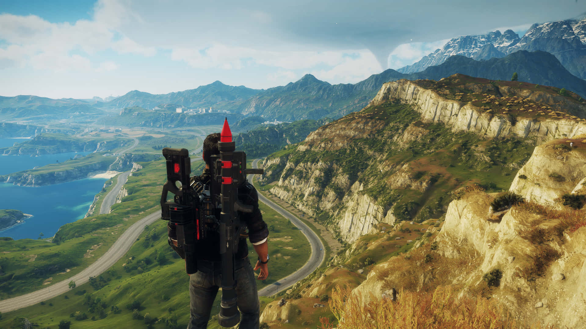 Just Cause 4 長路山景背景 壁紙 — 一個角色手持火箭發射器站在懸崖上，俯瞰蜿蜒的海岸公路和遠山，天空部分多雲。