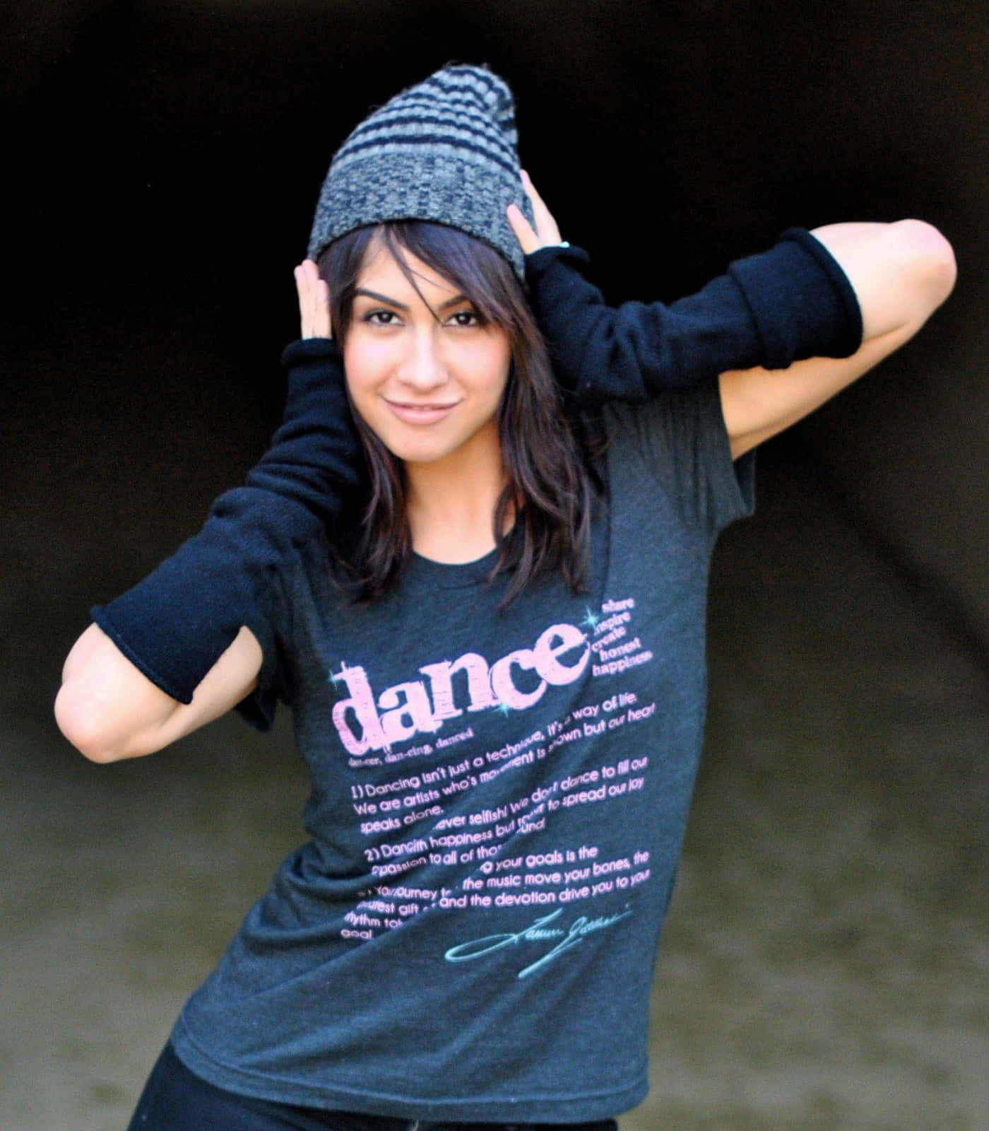 Just Dance 角色群舞 壁紙 — 一位微笑的女性，戴著灰色毛線帽和黑色襯衫，上面有舞蹈歌詞，雙手放在腦後，在深色背景前擺姿勢。