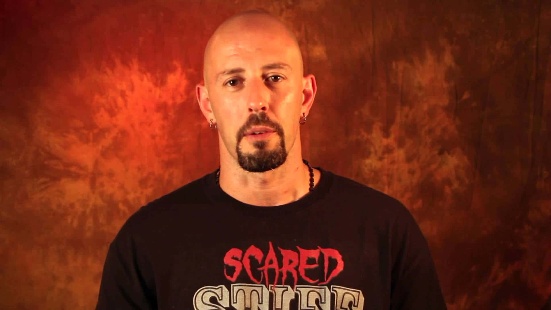 Justin Credible 紅色特寫 壁紙 — 一個光頭、留山羊鬍、戴耳環的男人，在火熱橙色紋理背景前直視鏡頭。