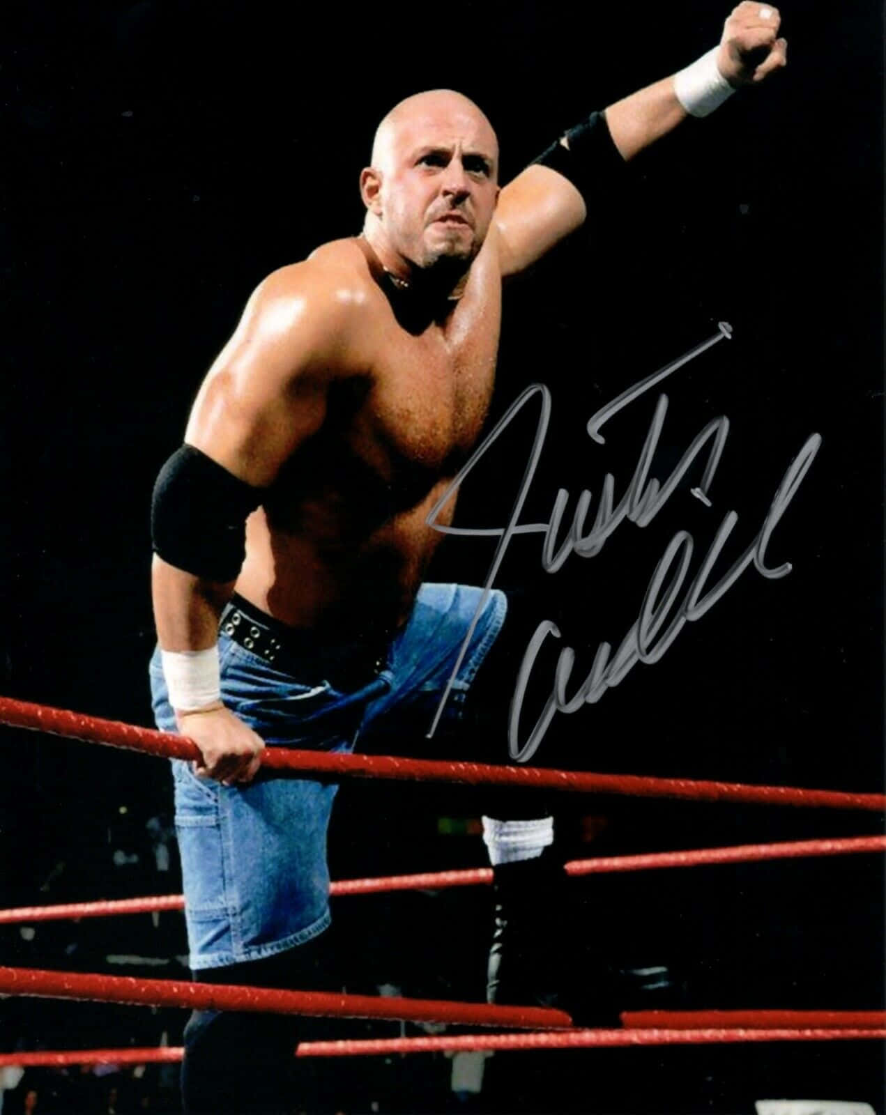 Justin Credible 簽名海報 壁紙 — 一個肌肉發達的光頭摔角手穿著牛仔短褲站在擂台上，在明亮的燈光下舉起拳頭，表情兇猛。