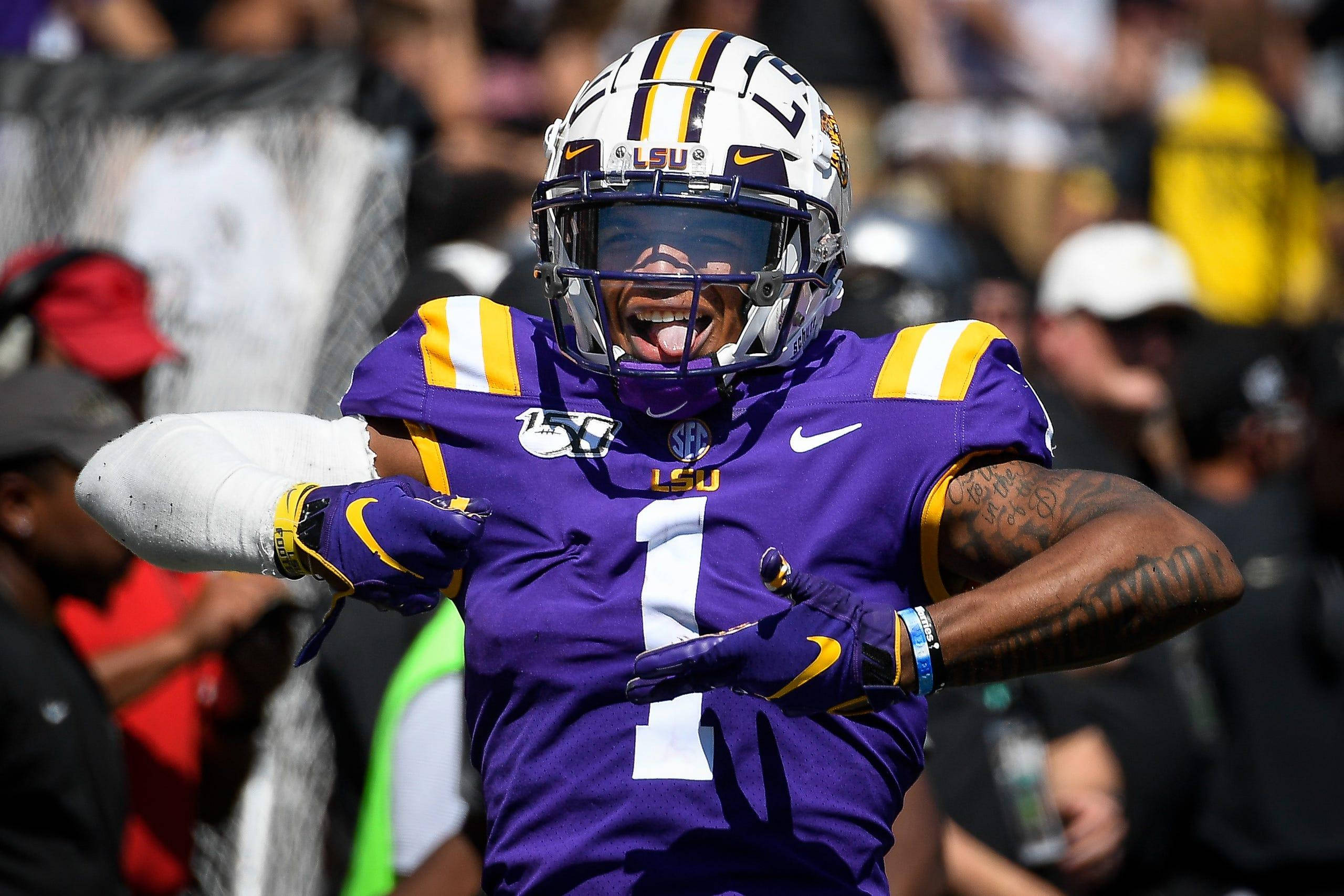 Justin Jefferson 身穿紫色球衣 壁紙 — 一位身穿紫色和金色裝備的LSU足球員在陽光明媚的比賽中張開雙臂、伸出舌頭慶祝。