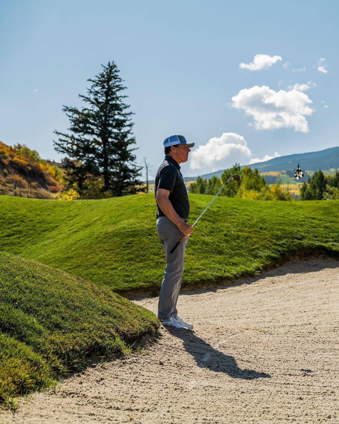 Justin Leonard 站立沙坑 壁紙 — 一位高爾夫球手站在陽光普照的沙坑中，俯瞰著由秋樹和遠山環繞的充滿活力的綠色球道。