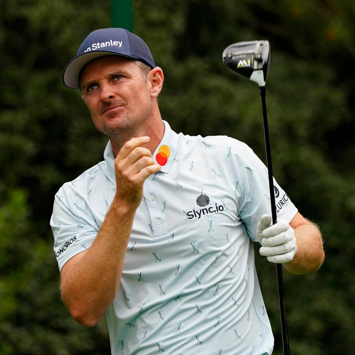 Justin Rose 擊球後凝視 壁紙 — 一位專注的高爾夫球手穿著淺藍色圖案襯衫和帽子，在模糊的綠色背景下拿著一號木桿並用硬幣做手勢。