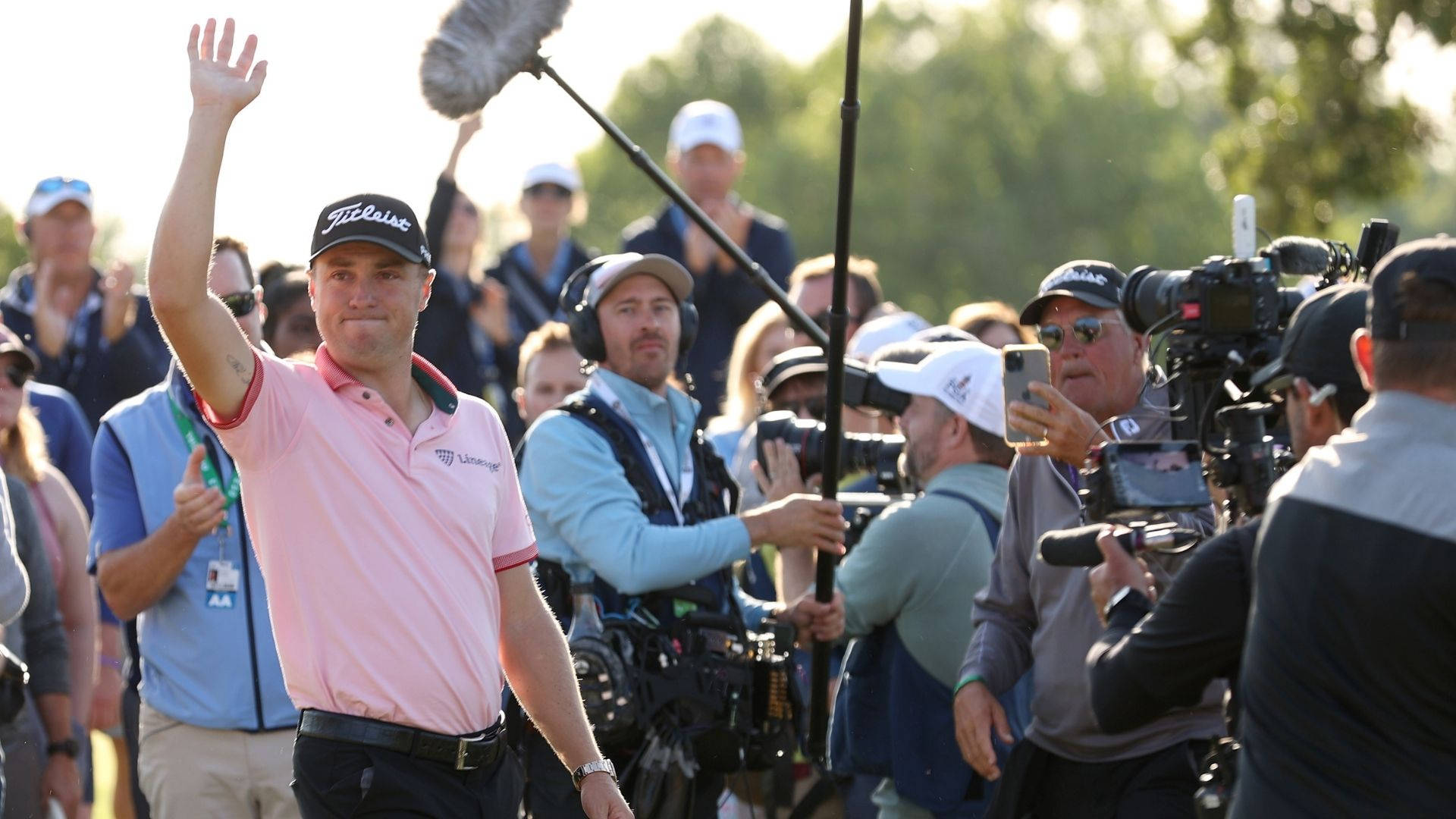 Justin Thomas 向觀眾揮手 壁紙 — 一名穿著粉紅色Polo衫的高爾夫球手在陽光明媚的高爾夫球場上向球迷揮手，周圍環繞著相機和工作人員。