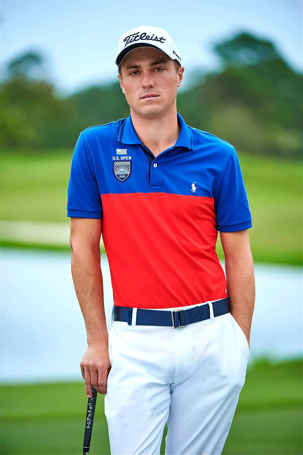 Justin Thomas 出現在美國公開賽 壁紙 — 一位專注的高爾夫球手身穿藍紅相間的Polo衫，站在翠綠的球場上，手持球桿，背景有樹木和水面。