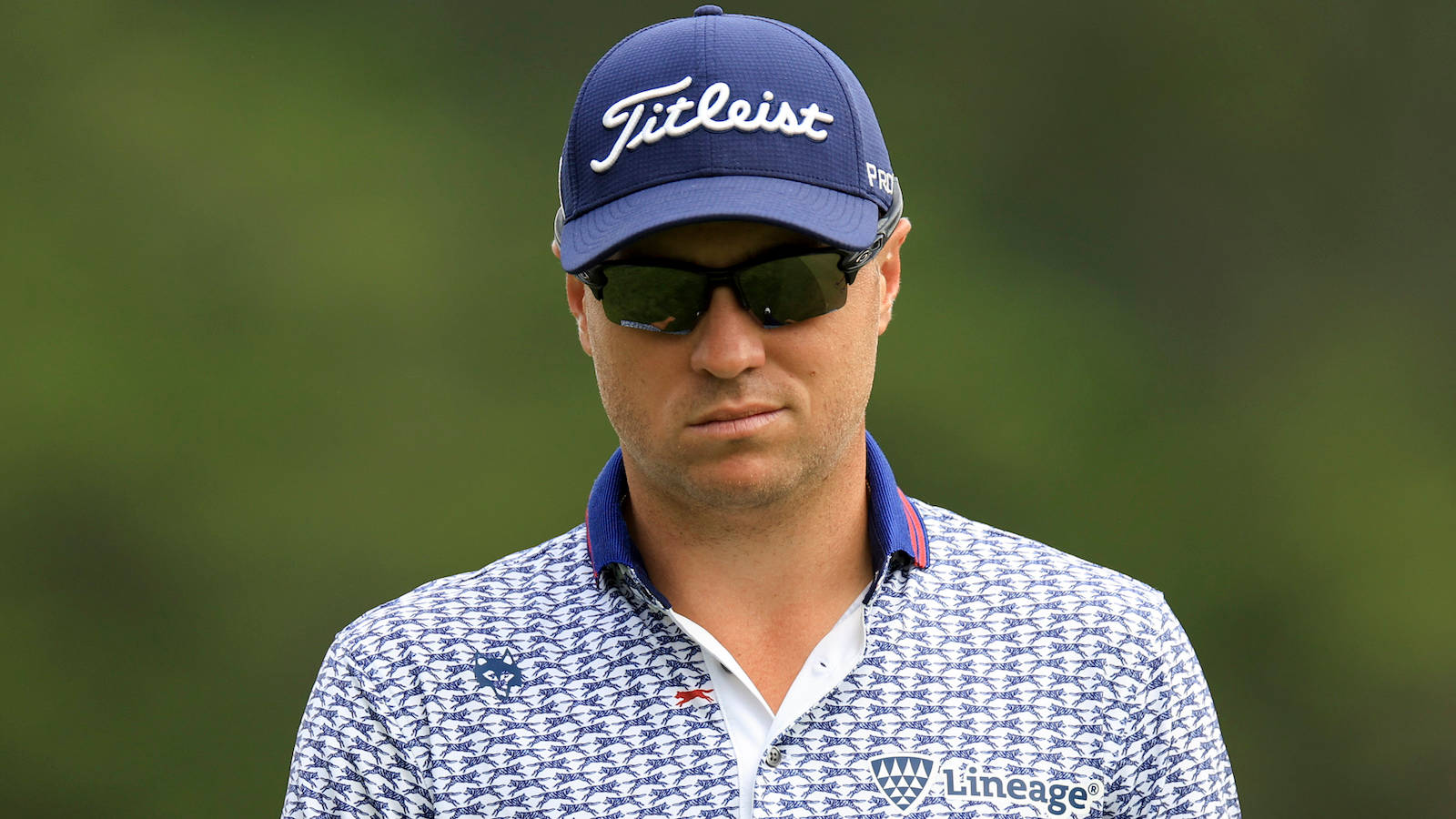 Justin Thomas 微笑挑眉 壁紙 — 一位專注的高爾夫球手，戴著藍色 Titleist 帽子和圖案 Polo 衫，站在模糊的綠色背景前。