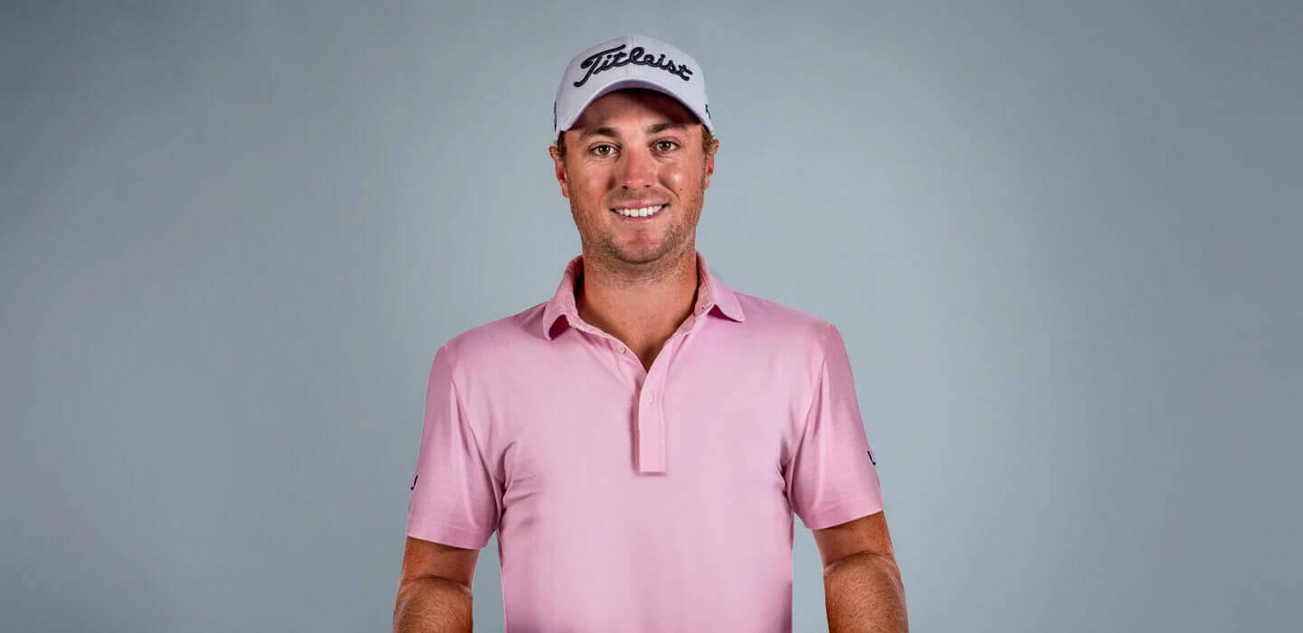 Justin Thomas 戴 Titleist 帽子 壁紙 — 一位微笑的高爾夫球手穿著粉紅色Polo衫和白色帽子，在純灰色工作室背景前擺姿勢。