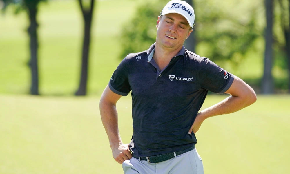 Justin Thomas 手叉腰的俏皮姿勢 壁紙 — 一位身穿海軍藍Polo衫和白色帽子、面帶微笑的高爾夫球手自信地站在陽光明媚的綠色高爾夫球場上，背景有樹木。