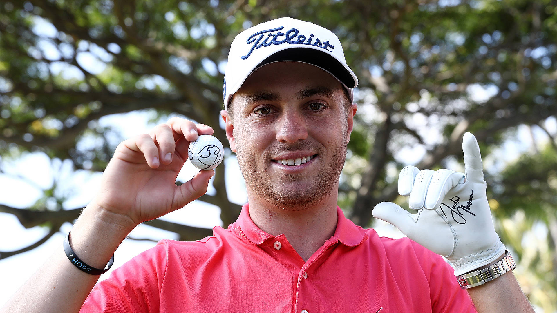 Justin Thomas 簽名高爾夫手套與球 壁紙 — 一位微笑的高爾夫球手穿著粉紅色Polo衫和白色帽子，拿著簽名高爾夫球，戴手套的手比出和平手勢。