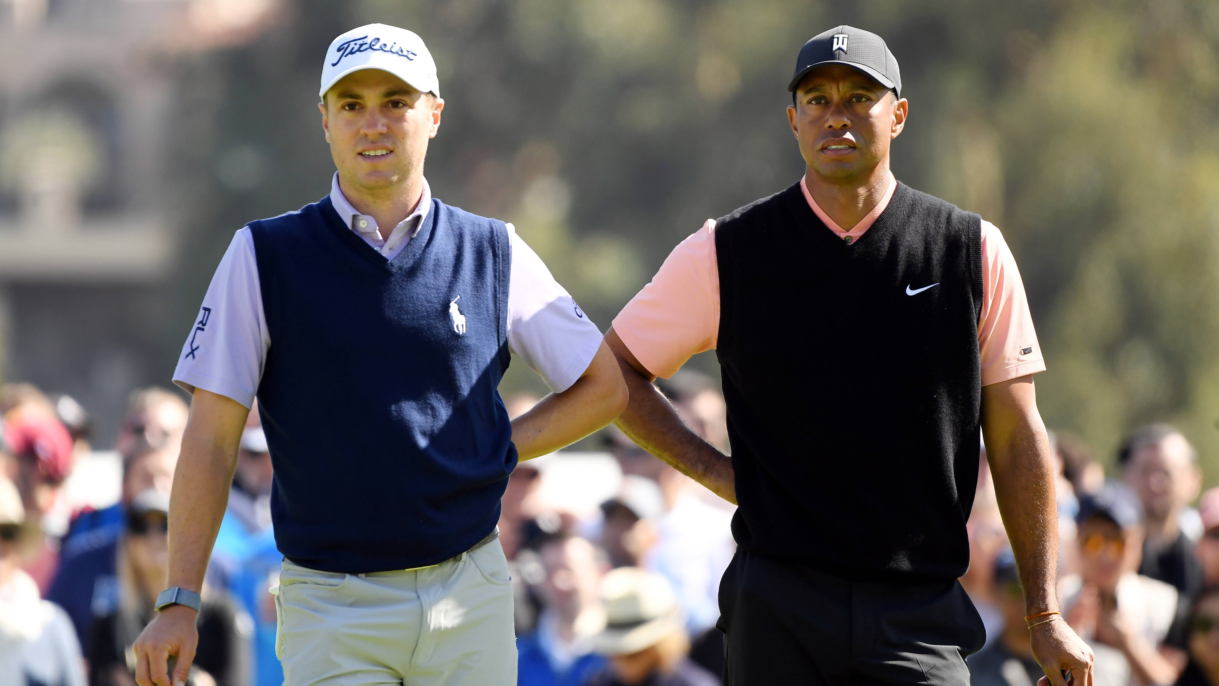 Justin Thomas 與 Tiger Woods 同框揮杆 壁紙 — 兩位職業高爾夫球手手挽手在陽光明媚的球場上行走，背景有觀眾。