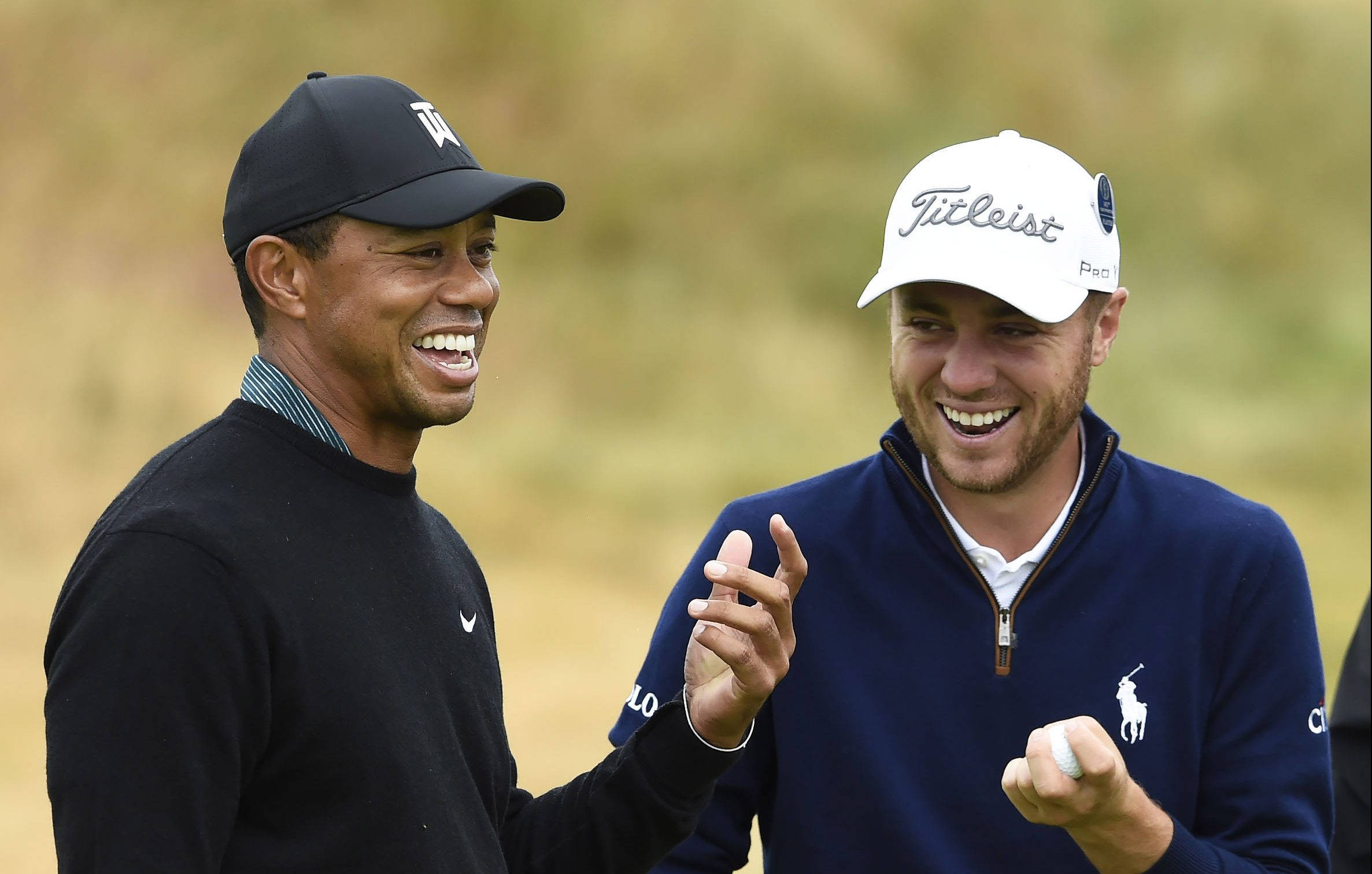 Justin Thomas 與 Tiger Woods 同歡 壁紙 — 兩名戴帽微笑的高爾夫球手在陽光明媚的球場上分享歡樂時刻，背景是模糊的草地。