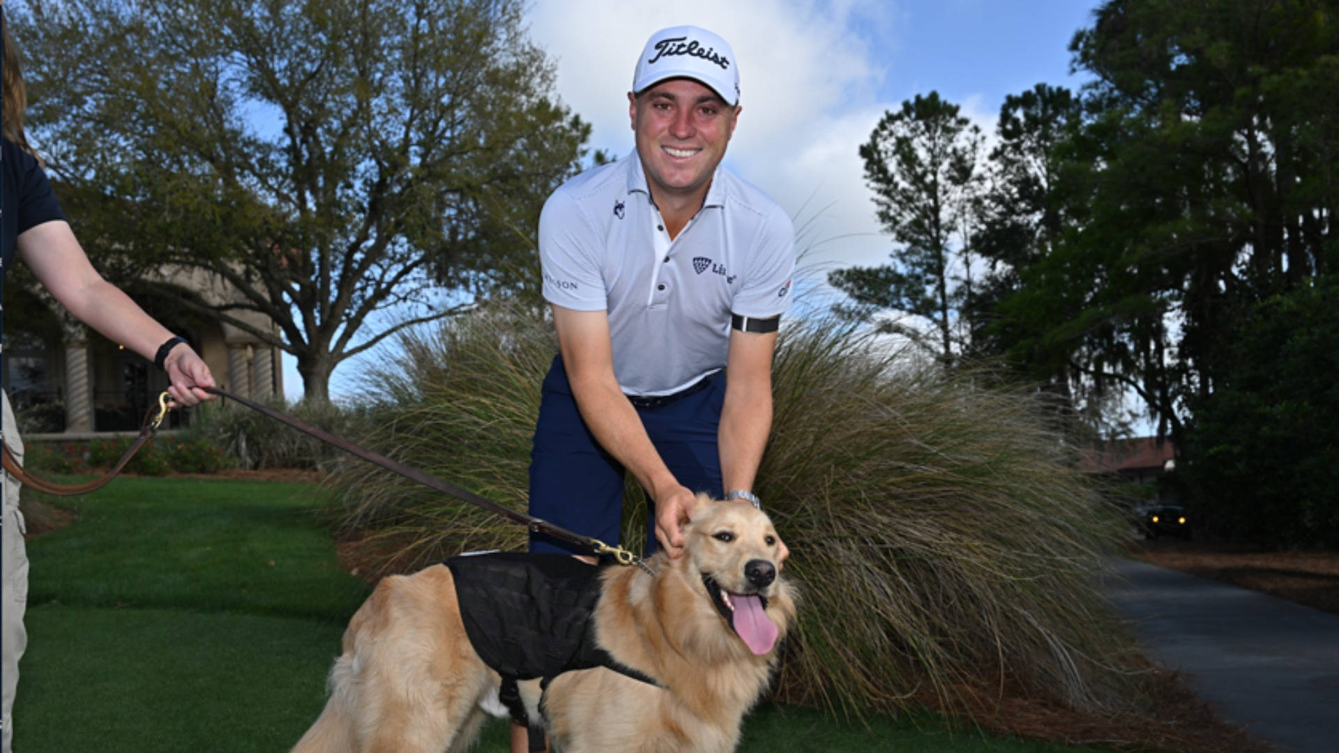 Justin Thomas 與金毛尋回犬 壁紙 — 一位微笑的高爾夫球手戴著白色帽子和Polo衫，彎腰撫摸一隻穿著黑色背心的金毛尋回犬，身後是草地、樹木和小徑。
