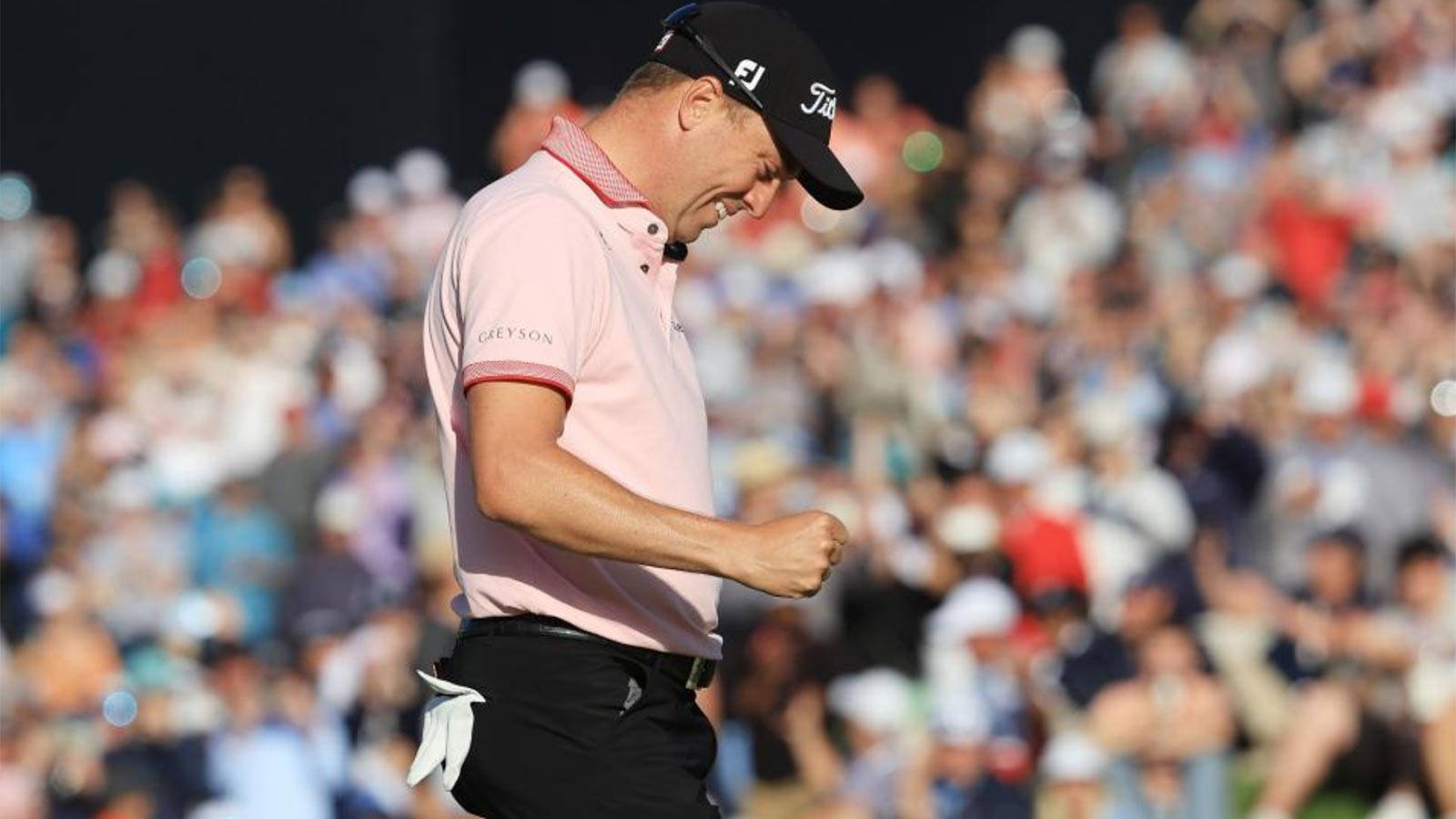 Justin Thomas 賽事擊球後慶祝 壁紙 — 一名身穿粉紅色Polo衫的高爾夫球手緊握拳頭慶祝勝利，身後模糊的人群為他歡呼。
