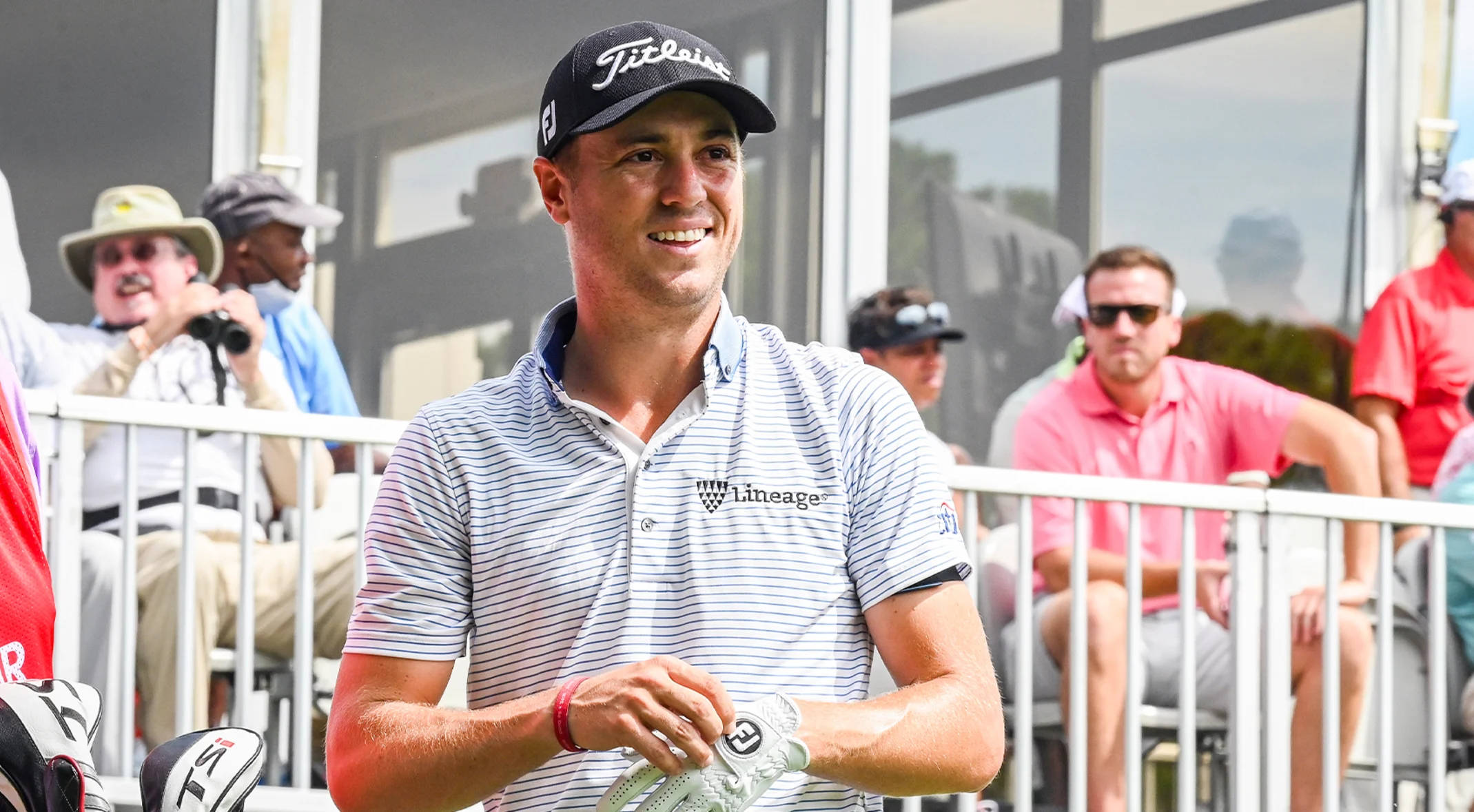 Justin Thomas 身穿條紋Polo衫參加高爾夫賽事 壁紙 — 一位微笑的高爾夫球手穿著條紋Polo衫和Titleist帽子，正在調整手套，觀眾在欄杆後觀看。