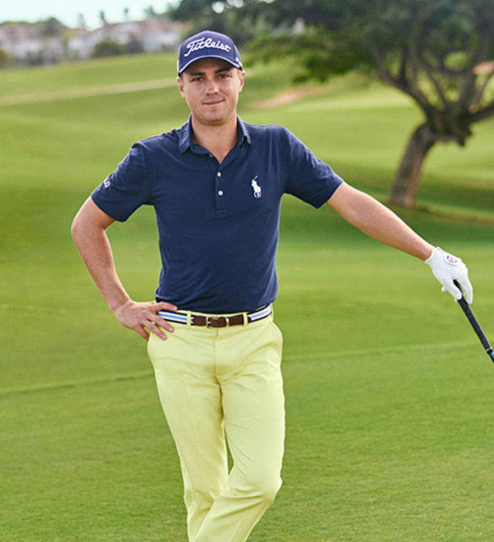 Justin Thomas 身穿黃色長褲 壁紙 — 一位自信的高爾夫球手身穿海軍藍Polo衫和黃色長褲，站在翠綠的球場上，手持球桿，背景有樹木。