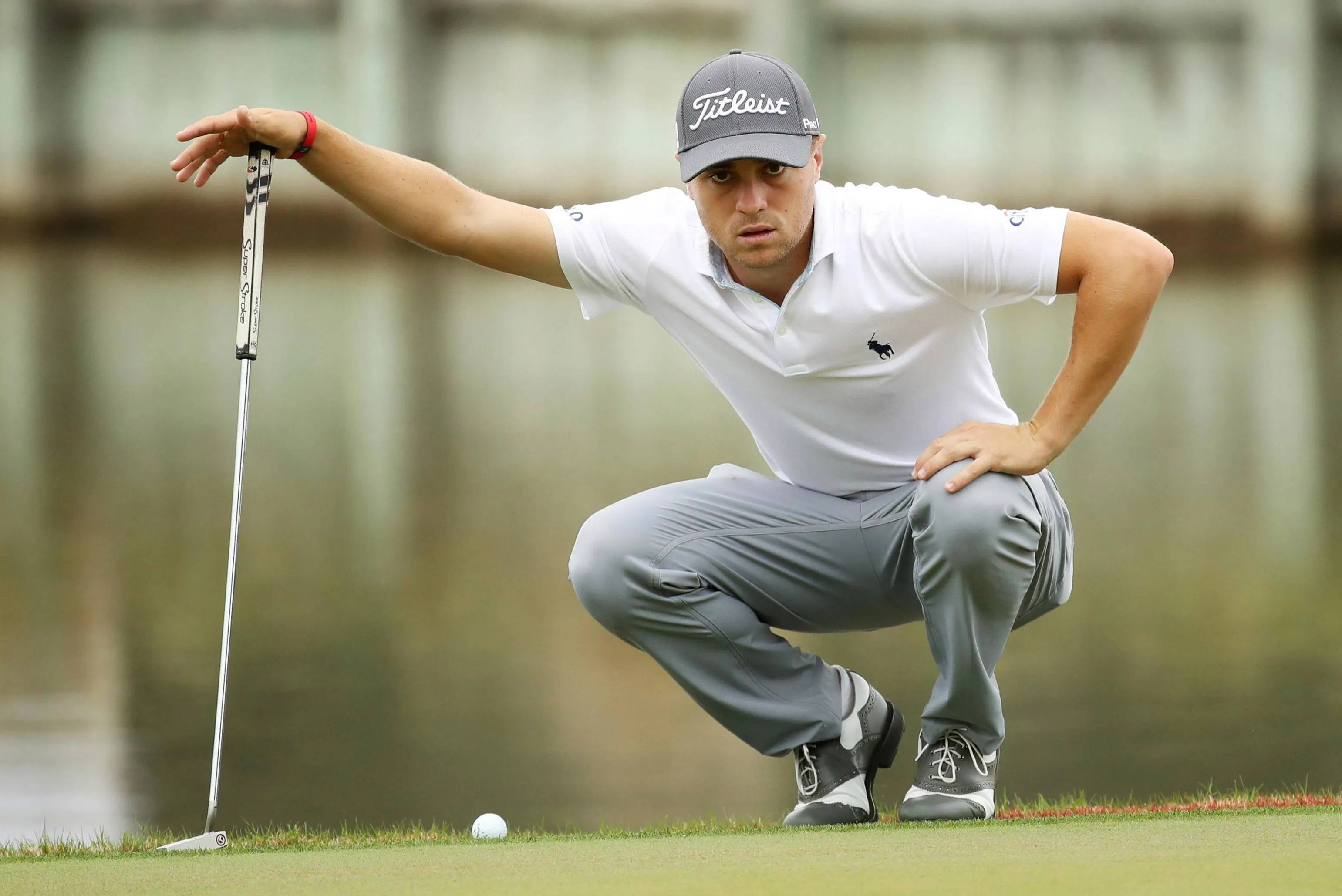 Justin Thomas 驚人凝視 壁紙 — 一位專注的高爾夫球手蹲在果嶺上，手持推桿靠近高爾夫球，分析他的推桿動作。